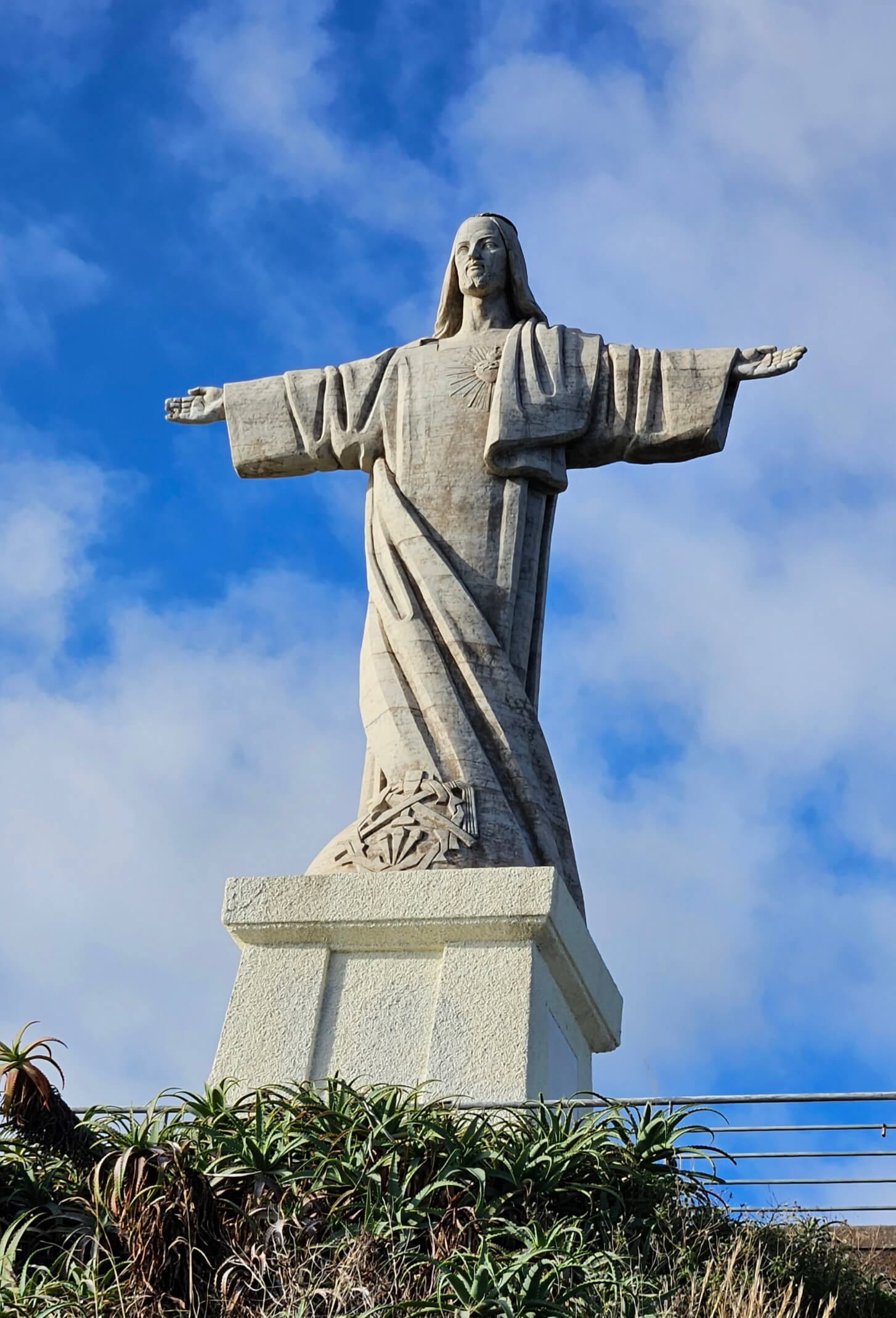 Eine große Statue von Christus mit ausgebreiteten Armen vor einem wolkigen Blauhimmel. Vegetation im Vordergrund.