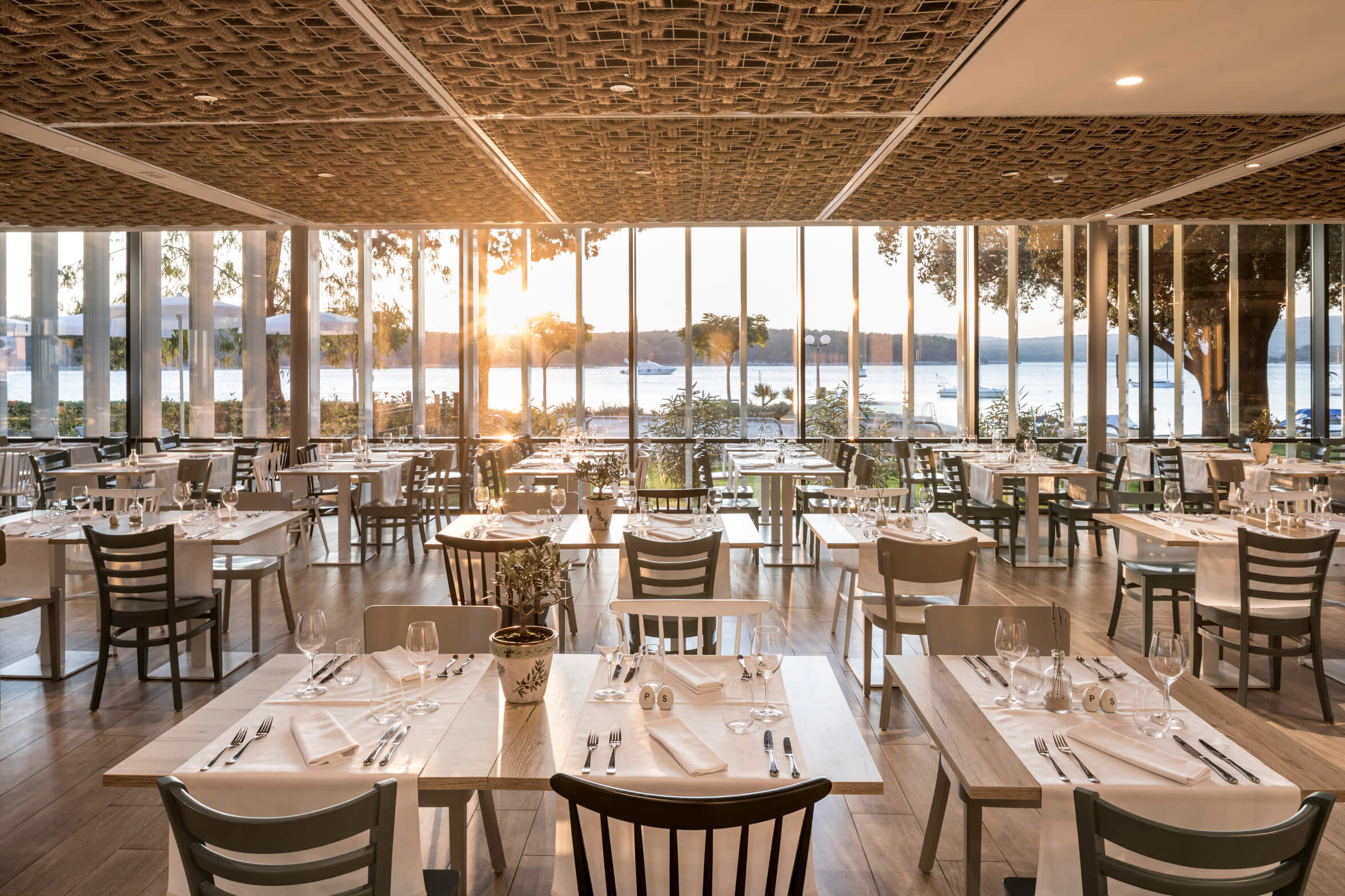 Ein modernes Restaurant mit Blick auf einen See, hell erleuchtet von Sonnenuntergang, elegant gedeckte Tische.