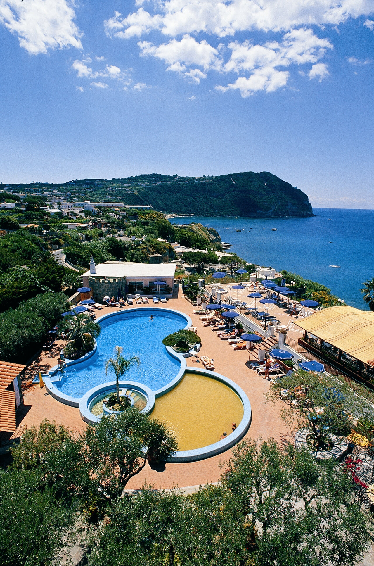 Schöne Poollandschaft mit Meerblick, umgeben von Grün und Liegen unter Sonnenschirmen an einem sonnigen Tag.