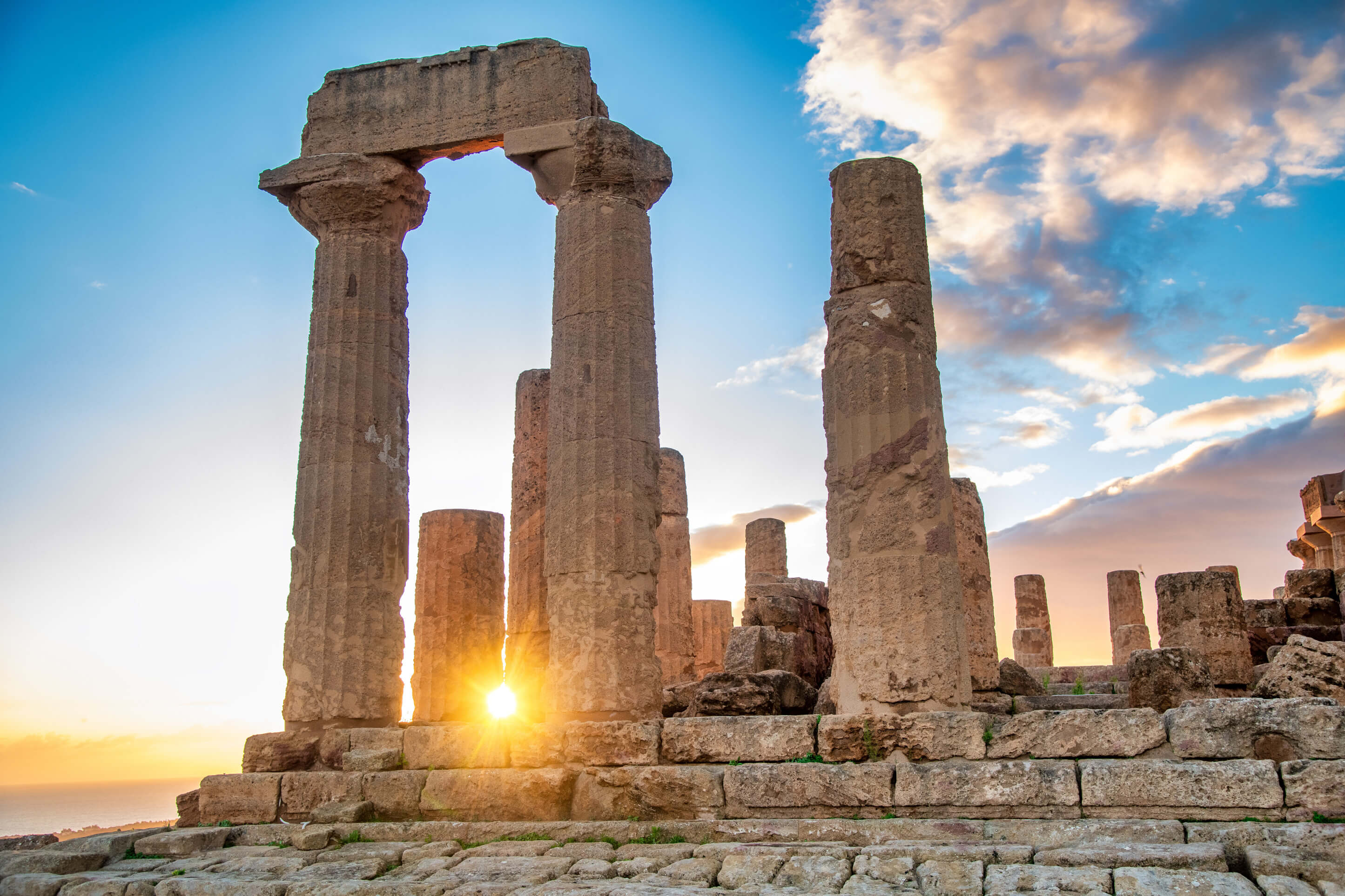 Antike Säulen stehen vor einem farbenfrohen Sonnenuntergang am Meer, unter einem klaren, blauen Himmel.