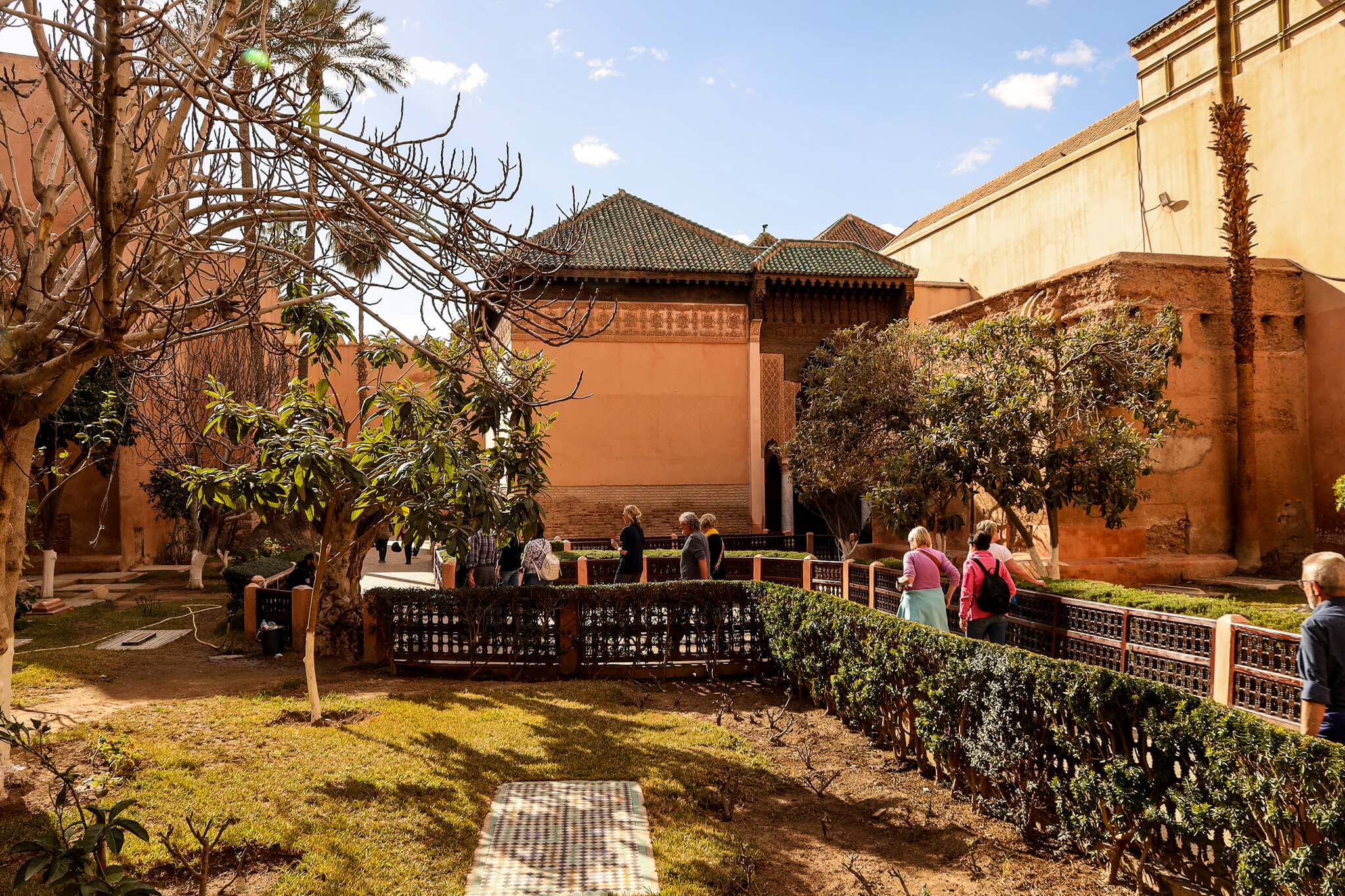 Ein Garten mit Bäumen und Besuchern vor einem historischen Gebäude, unter blauem Himmel in Marrakesch.