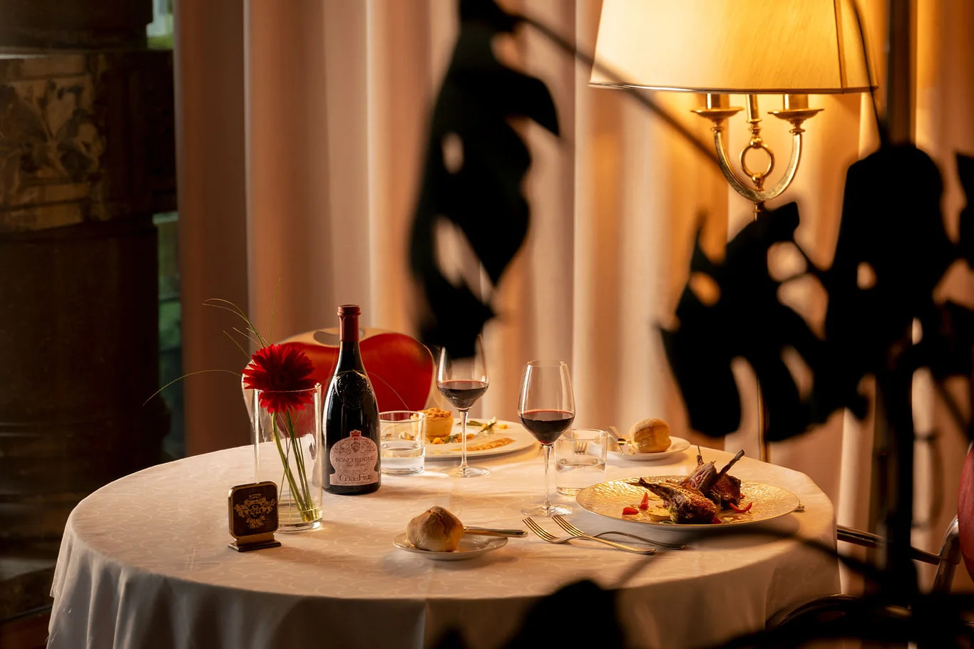 Elegant gedeckter Tisch mit Wein, köstlichem Essen und Blumen, beleuchtet von einer stilvollen Lampe.