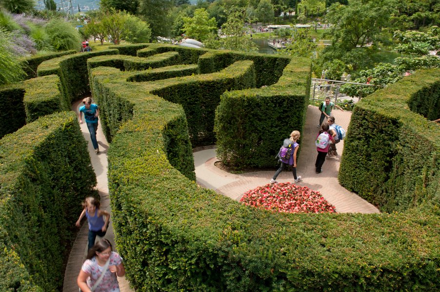 Ein labyrinthartiger Garten mit hohen Hecken, in dem Menschen umherlaufen und Blumen in der Mitte stehen.