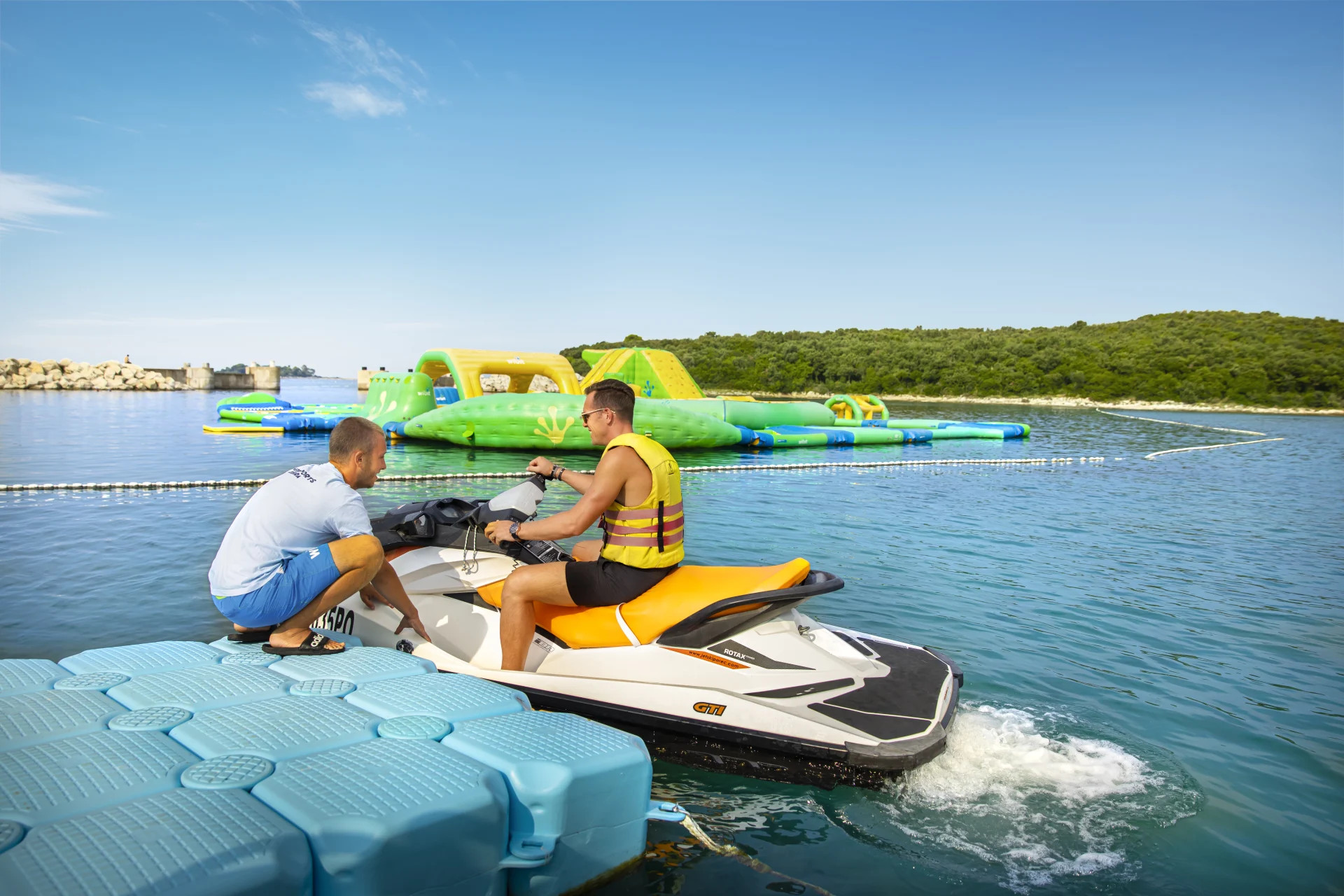Zwei Männer auf einem Jetski, im Hintergrund bunte Wasserattraktionen und eine klare blaue Lagune.