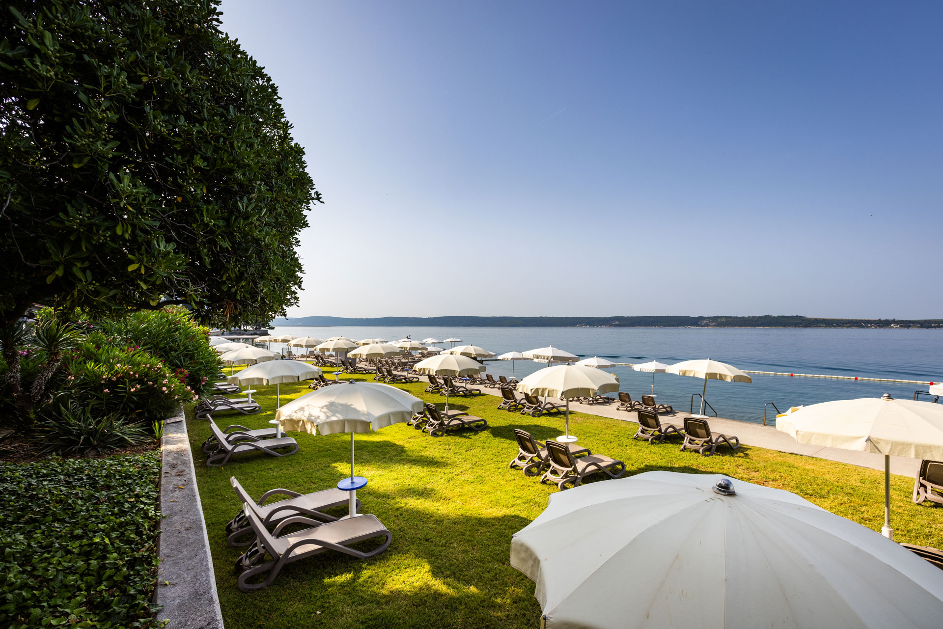 Strandliegestühle und Sonnenschirme am ruhigen Wasser, umgeben von grünem Rasen und Pflanzen, blauer Himmel.