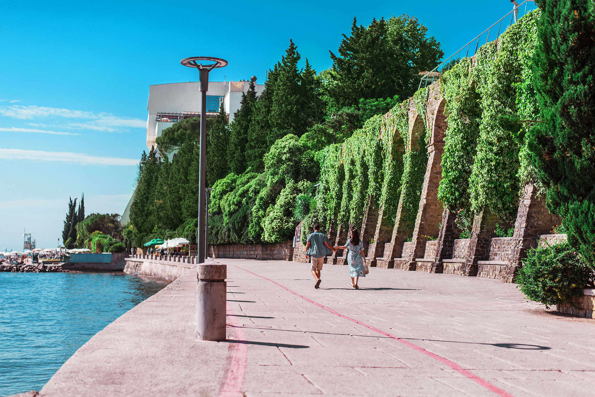 Ein Paar spaziert an einer sonnigen Uferpromenade, umgeben von üppigem Grün und einem blauen Himmel.