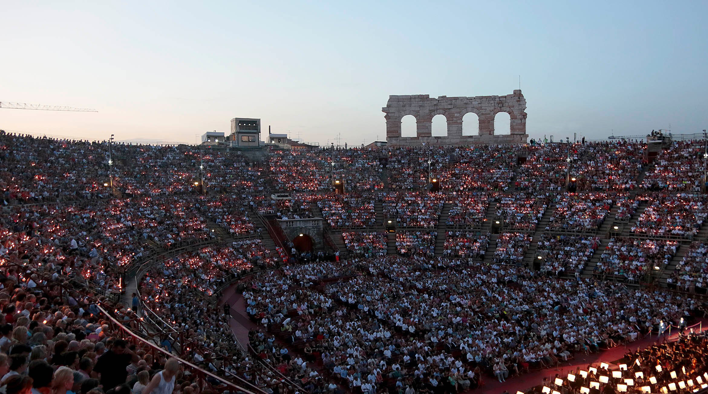 Vollbesetztes Amphitheater bei Sonnenuntergang, Besucher mit Kerzen, eine festliche Atmosphäre.
