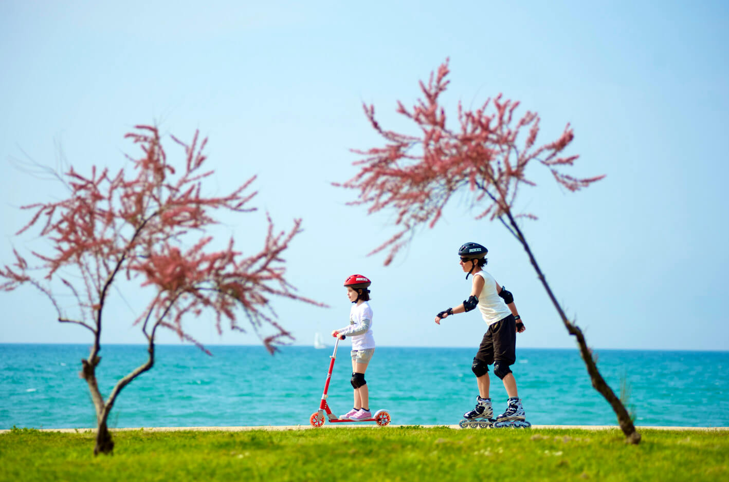 Zwei Kinder genießen einen sonnigen Tag am Strand: eines fährt Roller, das andere Rollschuhe mit schönem Ozeanhorizont.