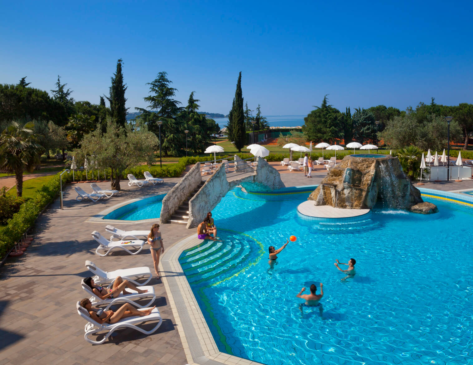 Ein sonniger Poolbereich mit spielenden Menschen, Liegen und Blick auf einen schönen Garten und das Meer.