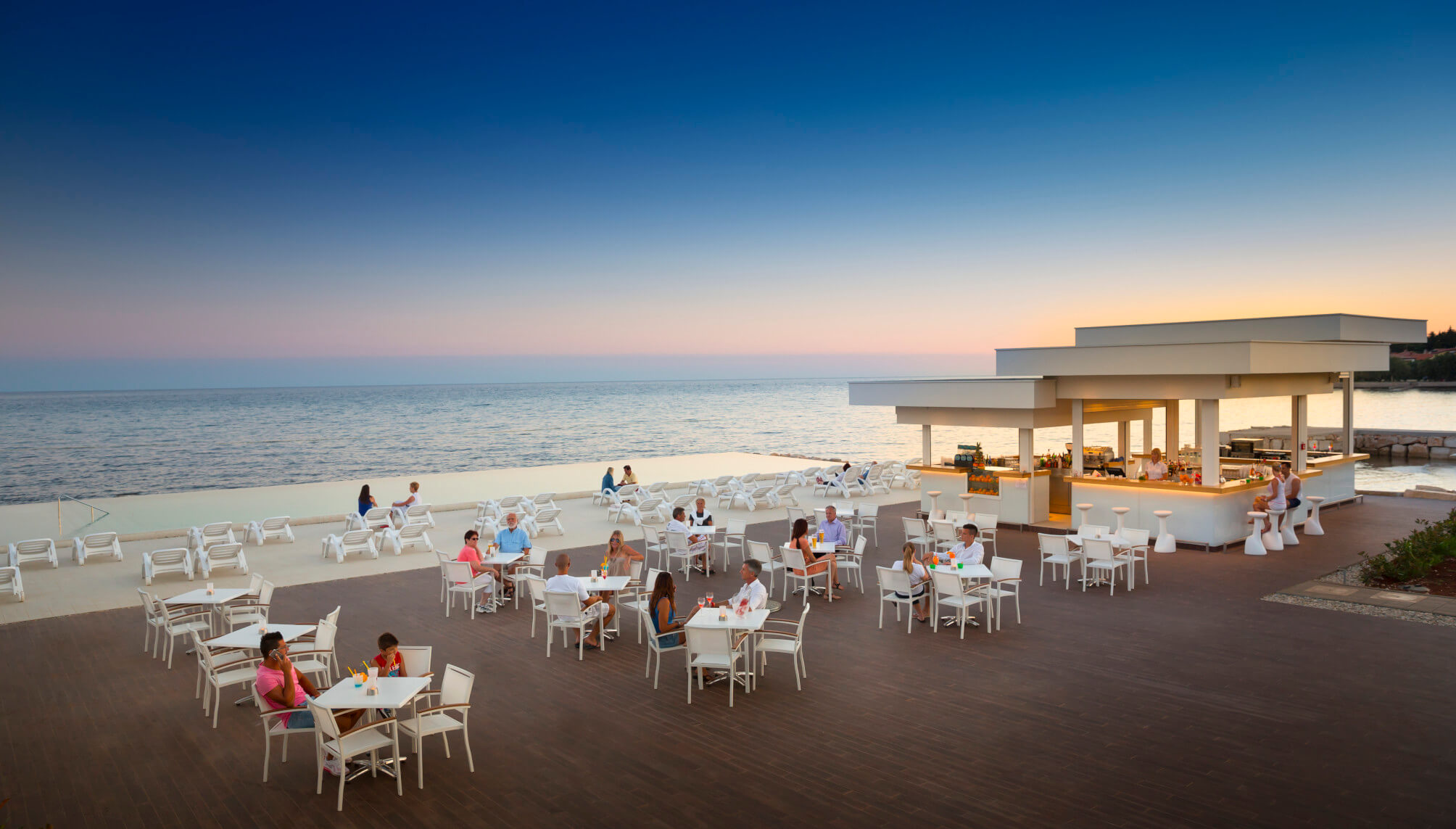 Ein stilvolles Restaurant am Meer, mit Gästen, die bei Sonnenuntergang entspannen und Getränke genießen.