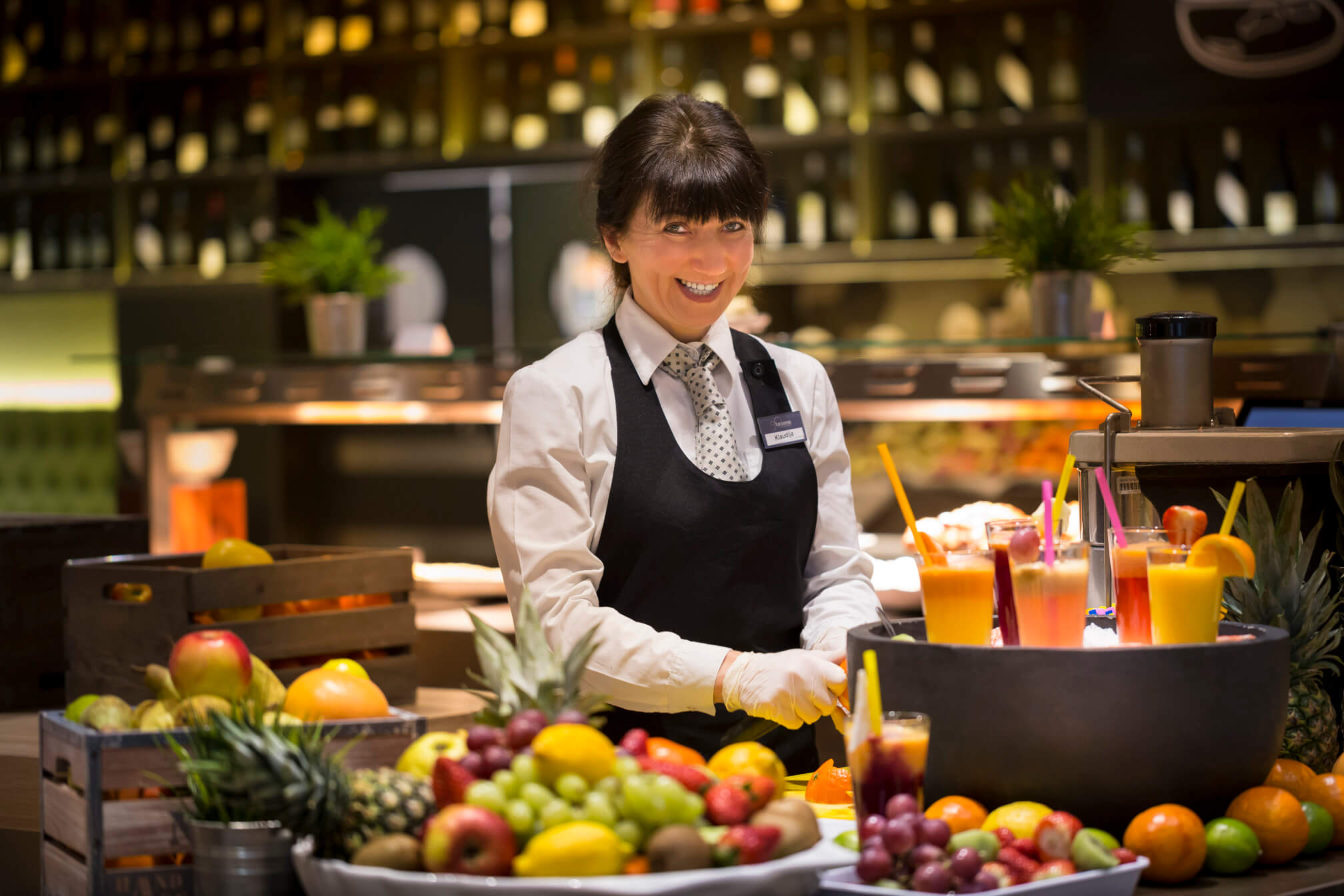Eine lächelnde Kellnerin in einem Restaurant mit frischen Früchten und bunten Cocktails im Hintergrund.
