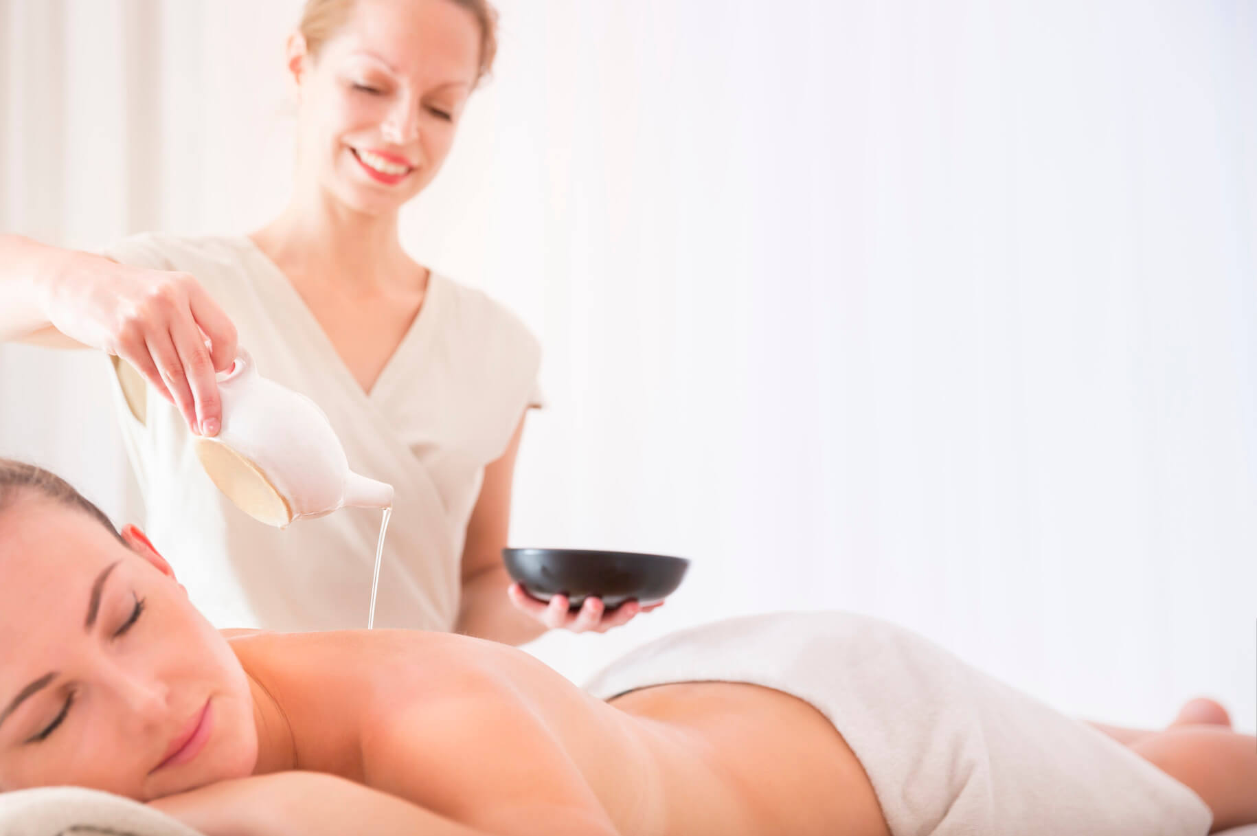 Eine entspannende Massage wird durchgeführt, während eine Frau Öl auf den Rücken einer liegenden Person gießt.