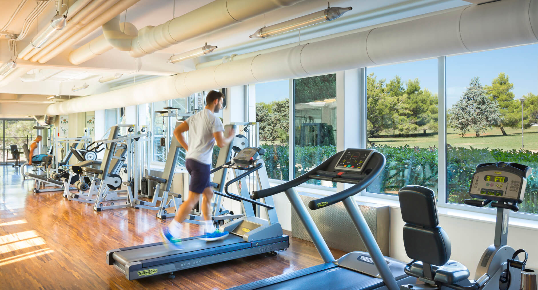 Ein moderner Fitnessraum mit Laufband und Kraftgeräten, große Fenster mit Blick auf die Natur.