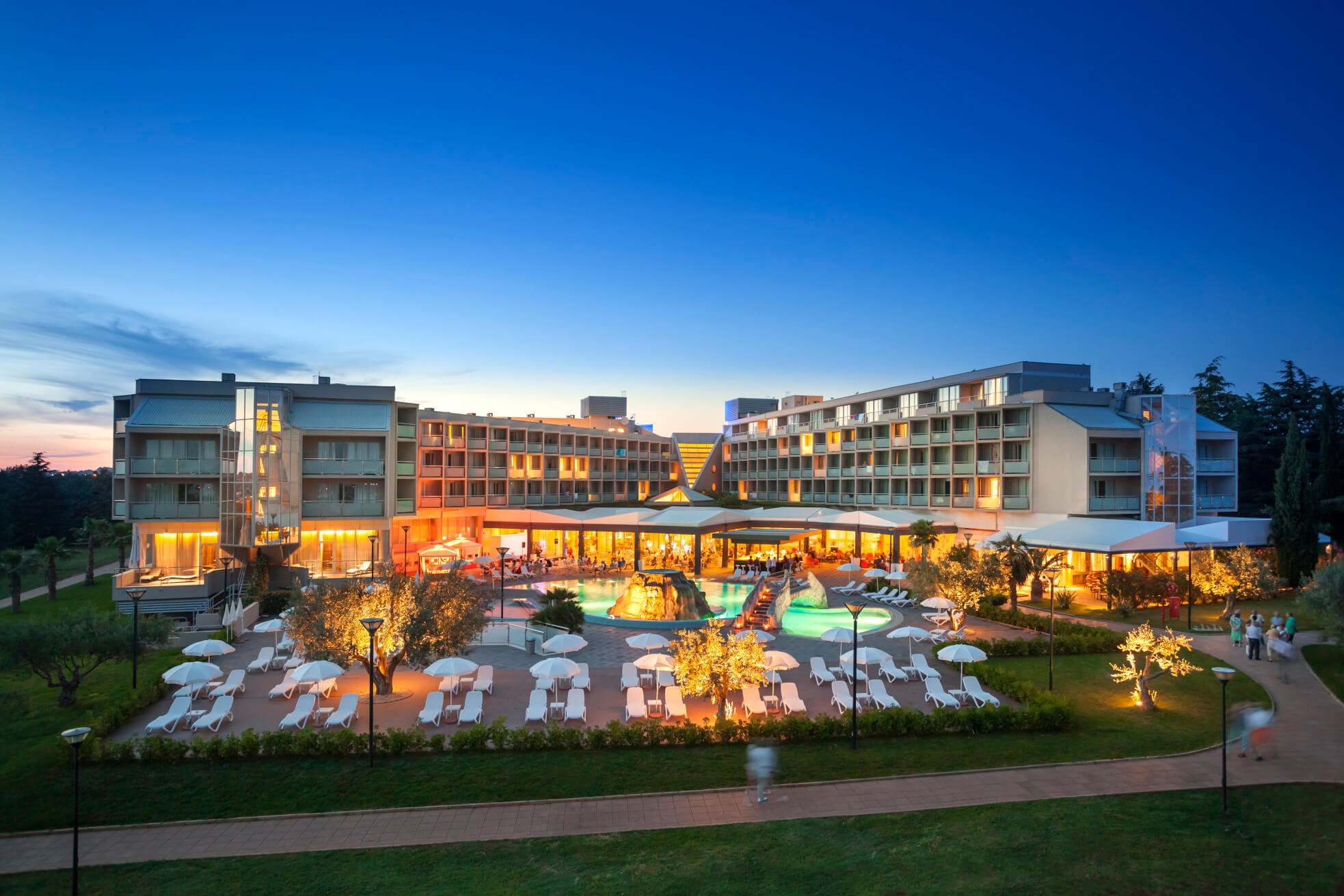 Modernes Hotel mit Pool und Lichtern in der Dämmerung, umgeben von einem gepflegten Garten und Sonnenliegen.