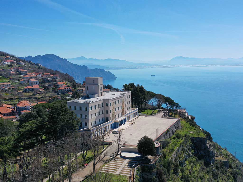 Ein modernes Gebäude liegt an einer Küste, umgeben von Bergen und blauen Meer, unter strahlend blauem Himmel.