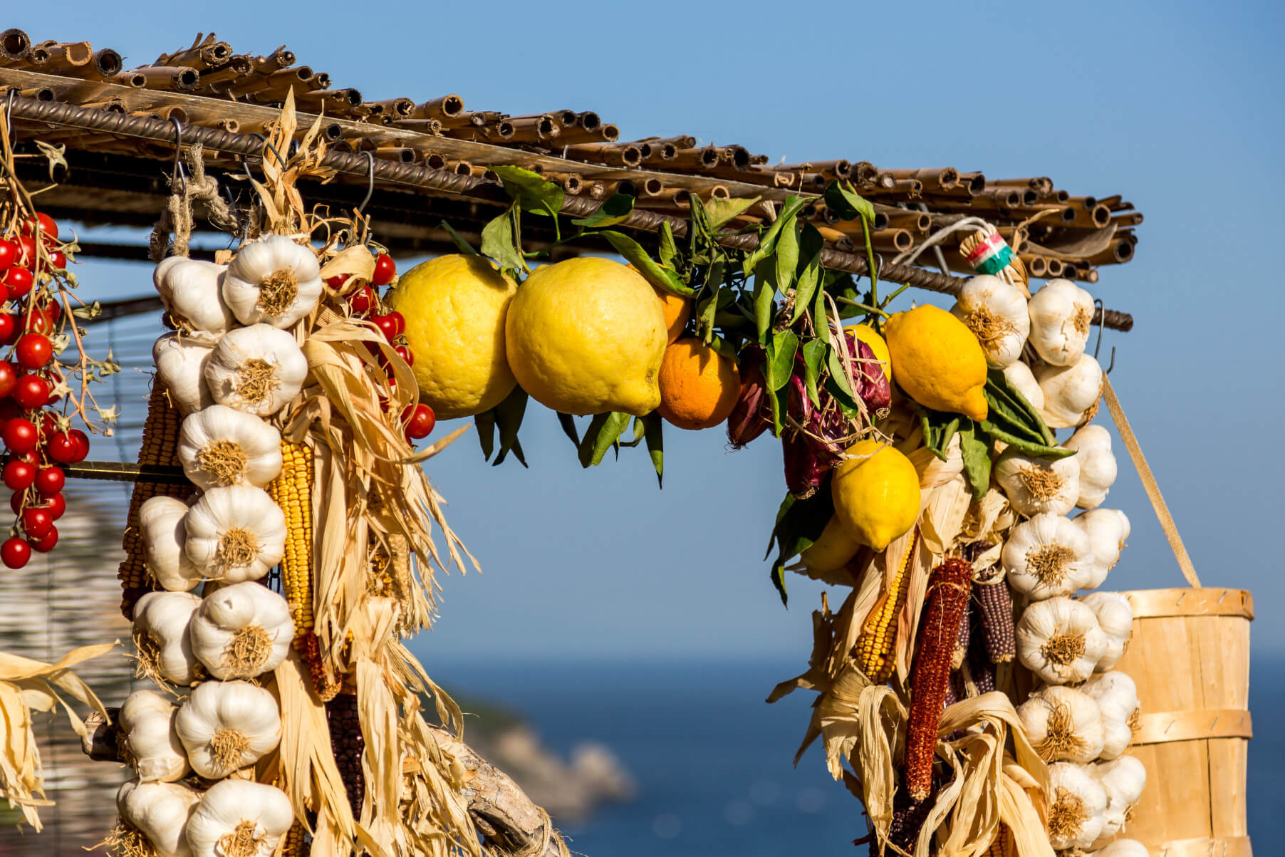Bunte Früchte und Knoblauch hängen exotisch dekoriert unter einem Dach mit Meeresblick im Hintergrund.