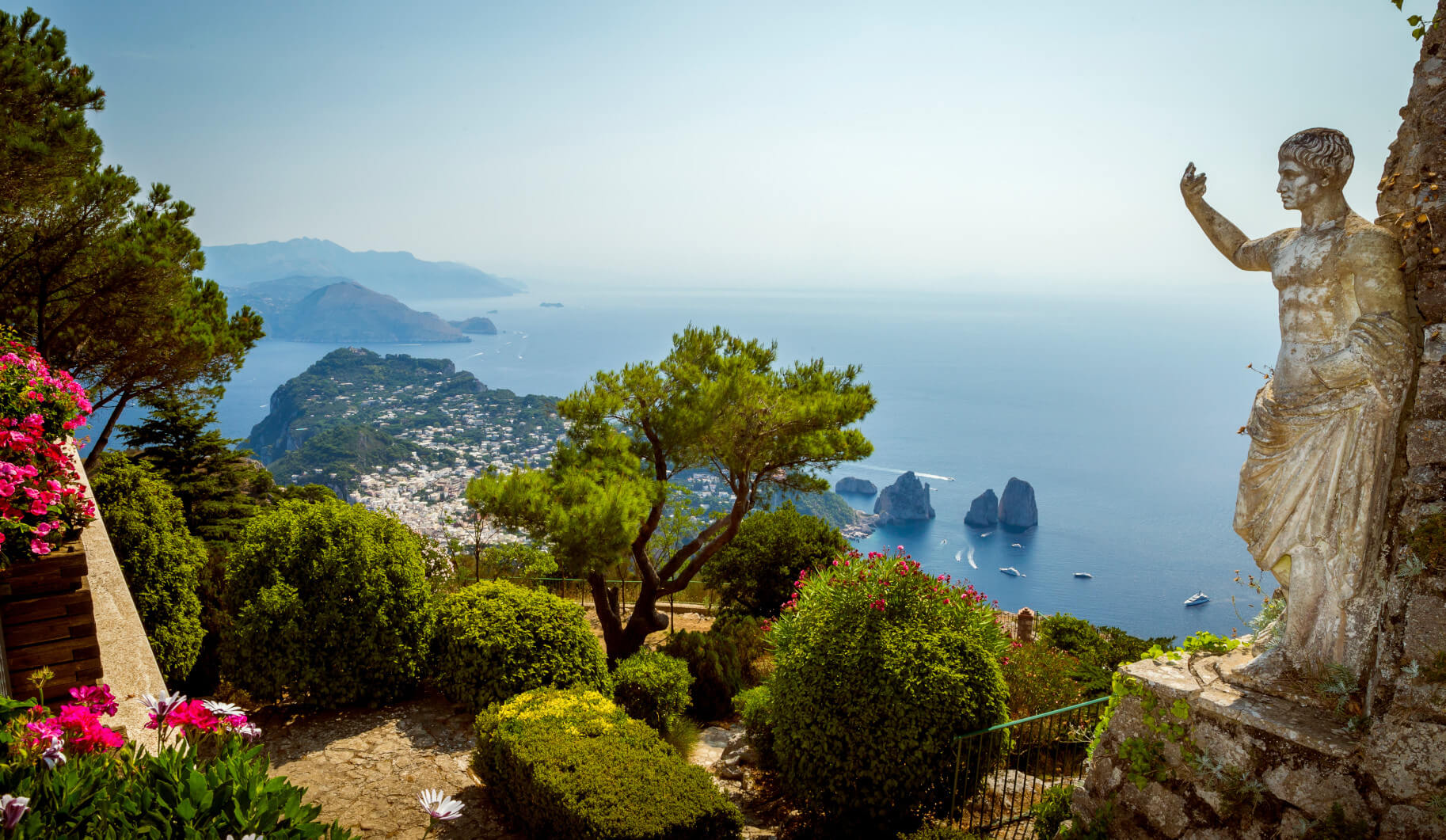 Blick auf die Küste von Capri mit einer Statue, üppigen Pflanzen und dem Meer im Hintergrund.