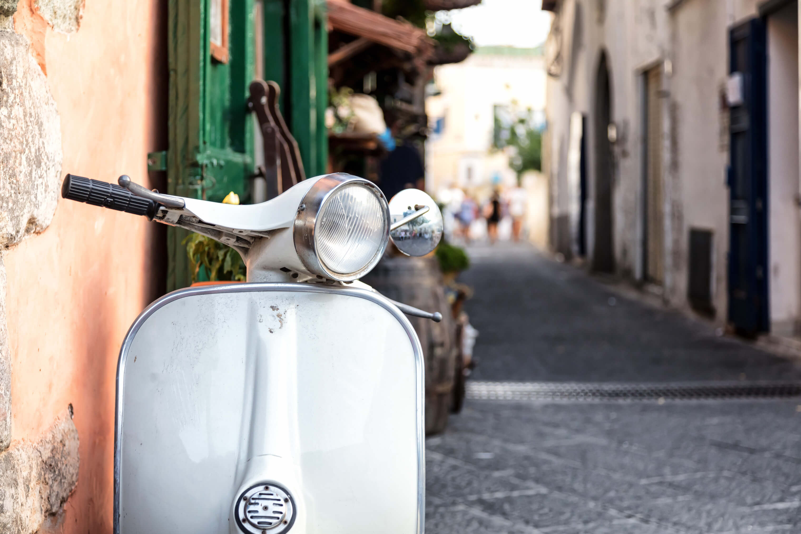 Ein weißer Roller steht in einer malerischen Gasse mit alten Gebäuden und unscharfen Menschen im Hintergrund.