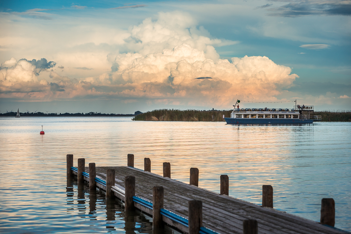 Ein Steg führt zu einem ruhigen See, wo ein Boot und beeindruckende Wolken zu sehen sind. Eine friedliche Szene.