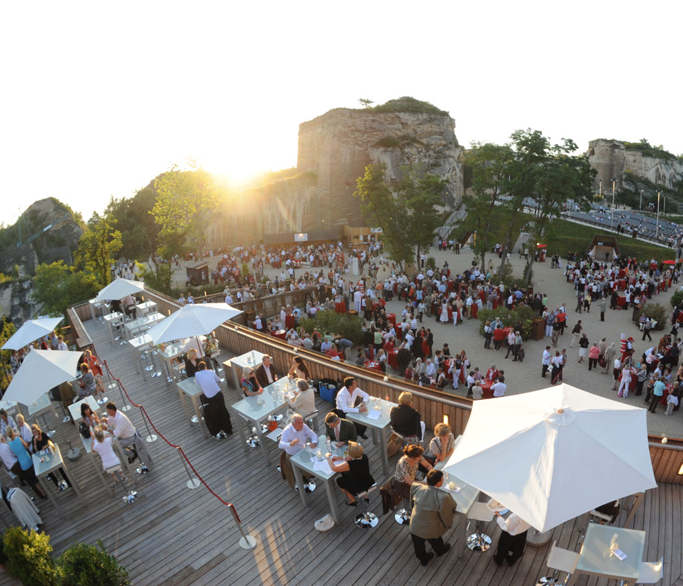 Eine lebhafte Veranstaltung im Freien, mit vielen Menschen, Tischen und Sonnenschirmen vor einer schönen Kulisse.