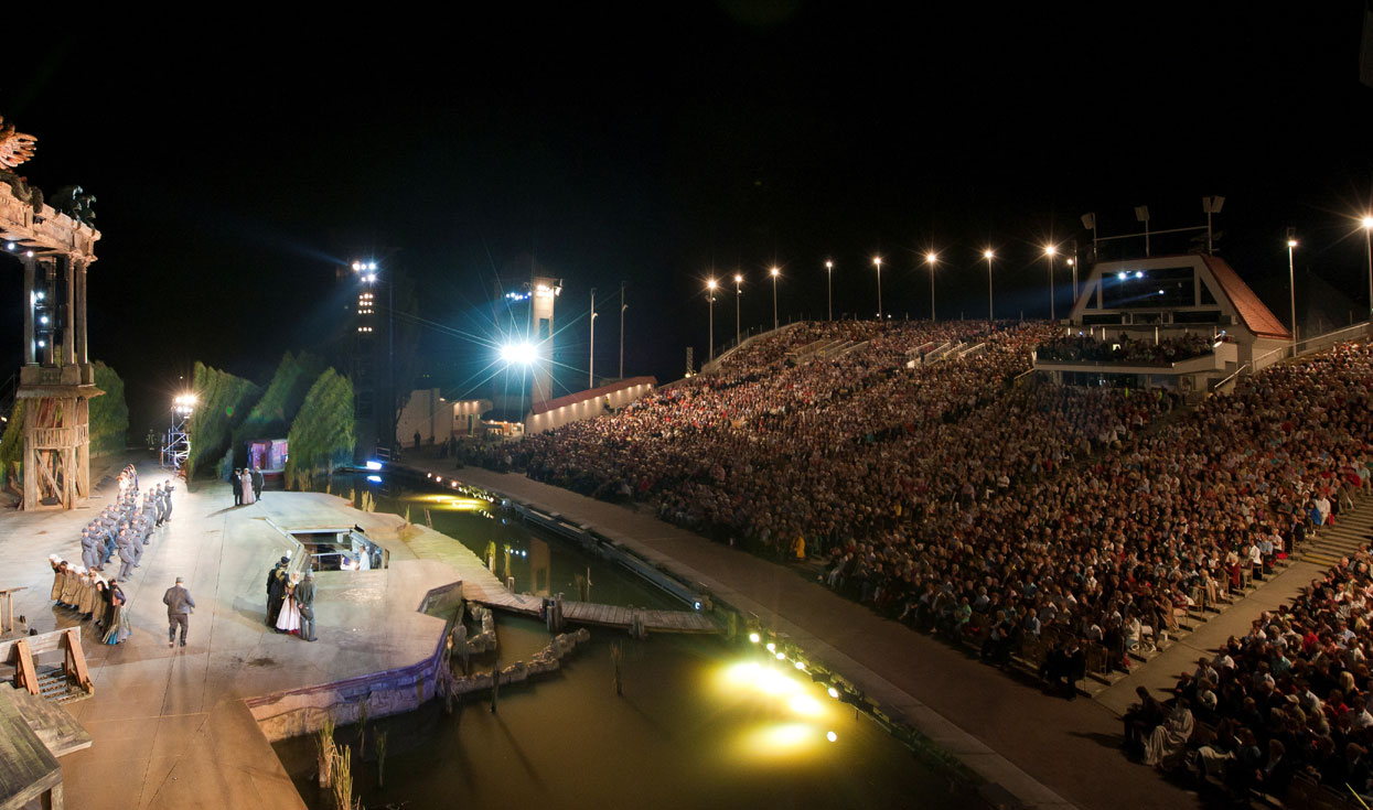 Eine große Menschenmenge verfolgt eine Abendaufführung auf einer Freiluftbühne bei Nacht.