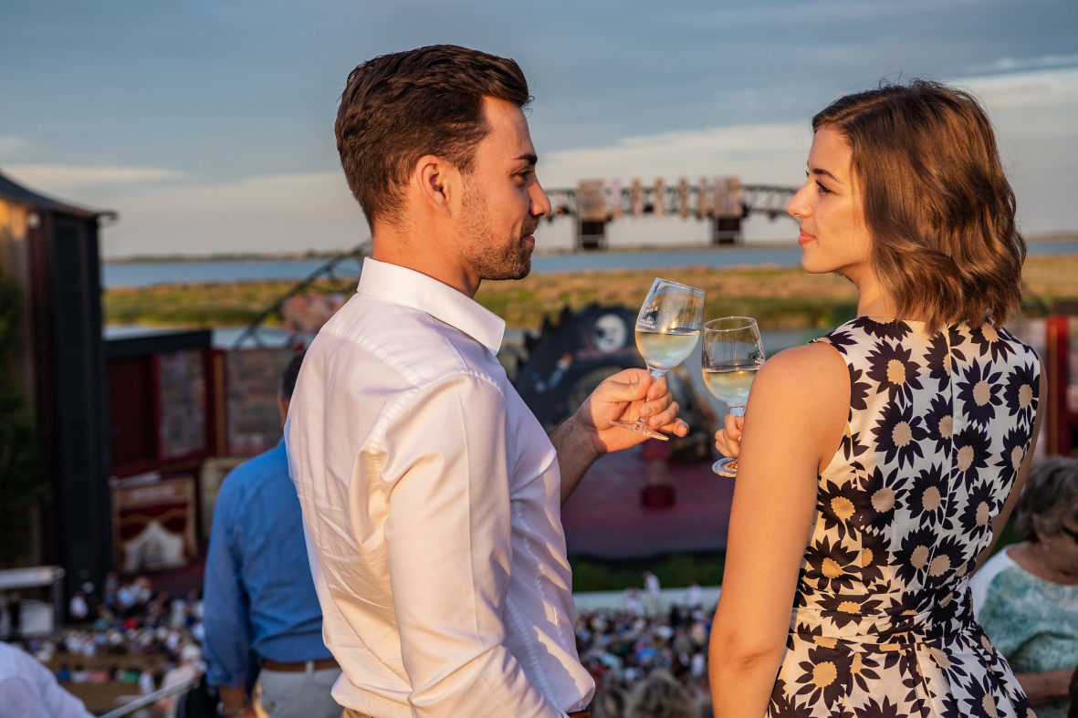 Ein Paar prostet sich mit Weingläsern zu, während im Hintergrund eine Aufführung stattfindet.