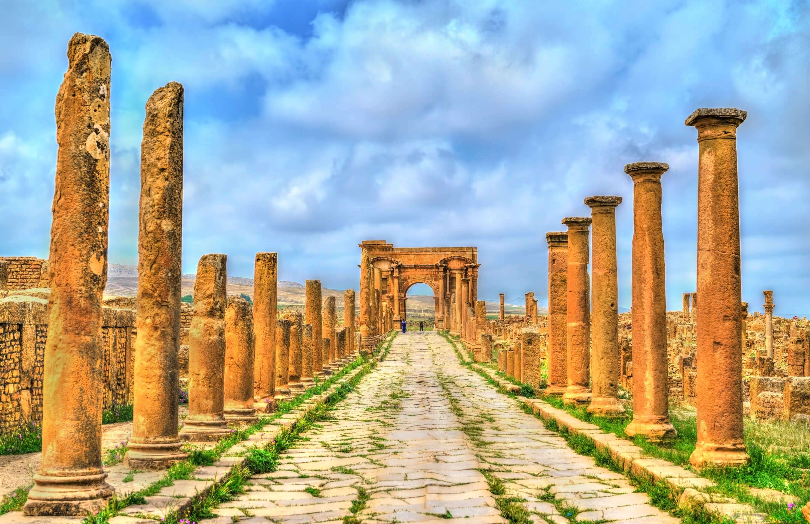 Eine antike Straße mit Säulen und einem Bogen, umgeben von einer dramatischen Wolkenlandschaft.