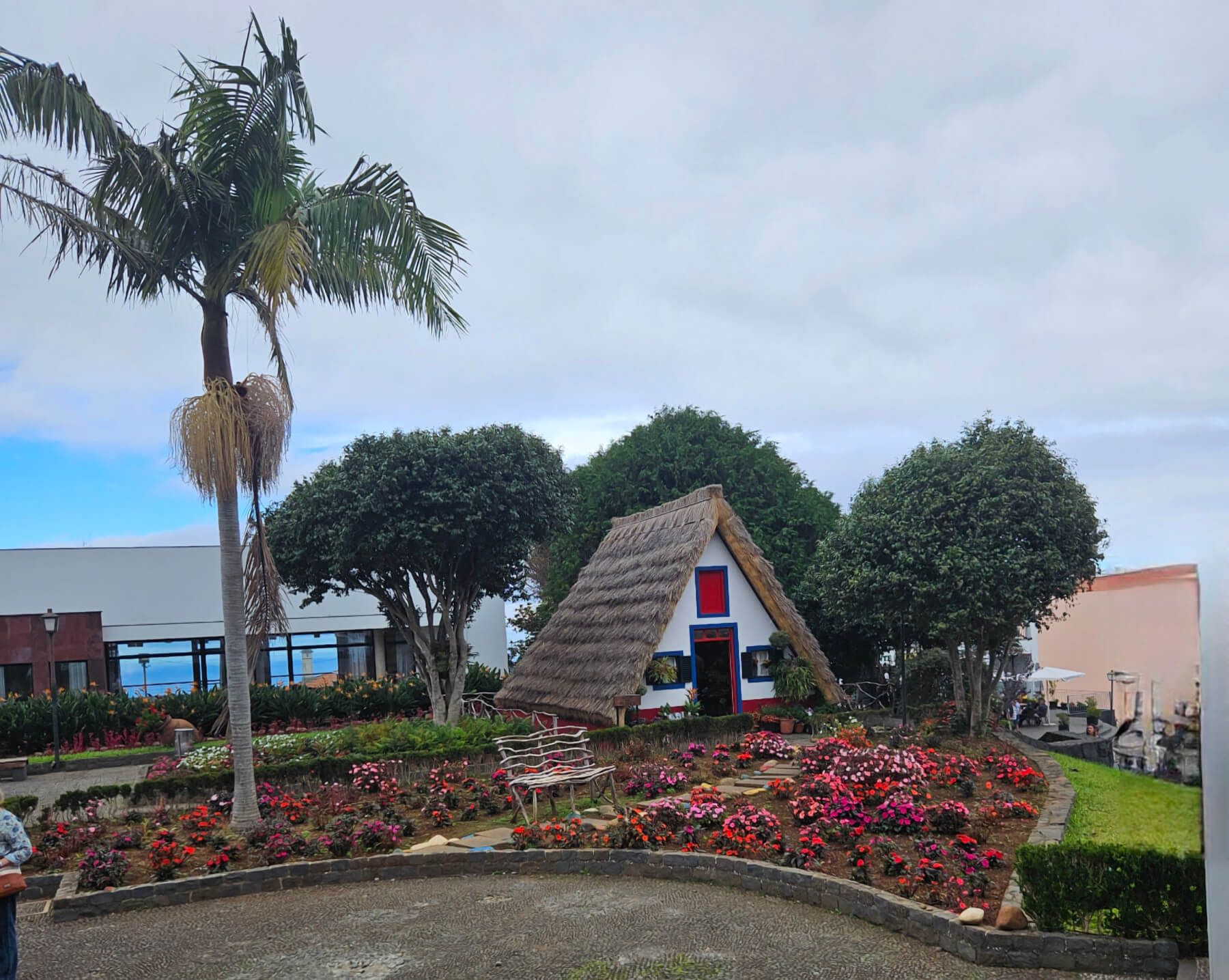 Ein bunter Garten mit einer kleinen, strohgedeckten Hütte und Palmen umgeben von blühenden Blumen.