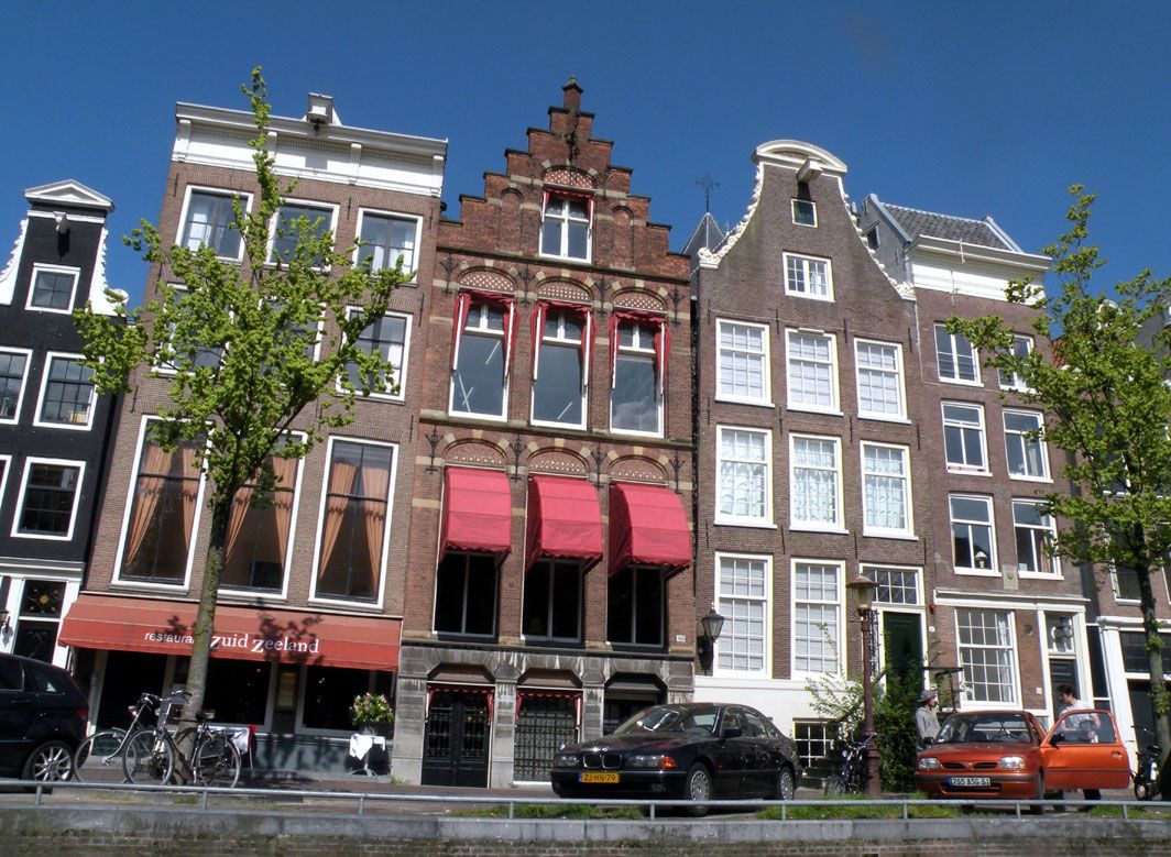 Bunte, historische Gebäude in Amsterdam mit roten Markisen und Bäumen vor der Fassade an einem sonnigen Tag.