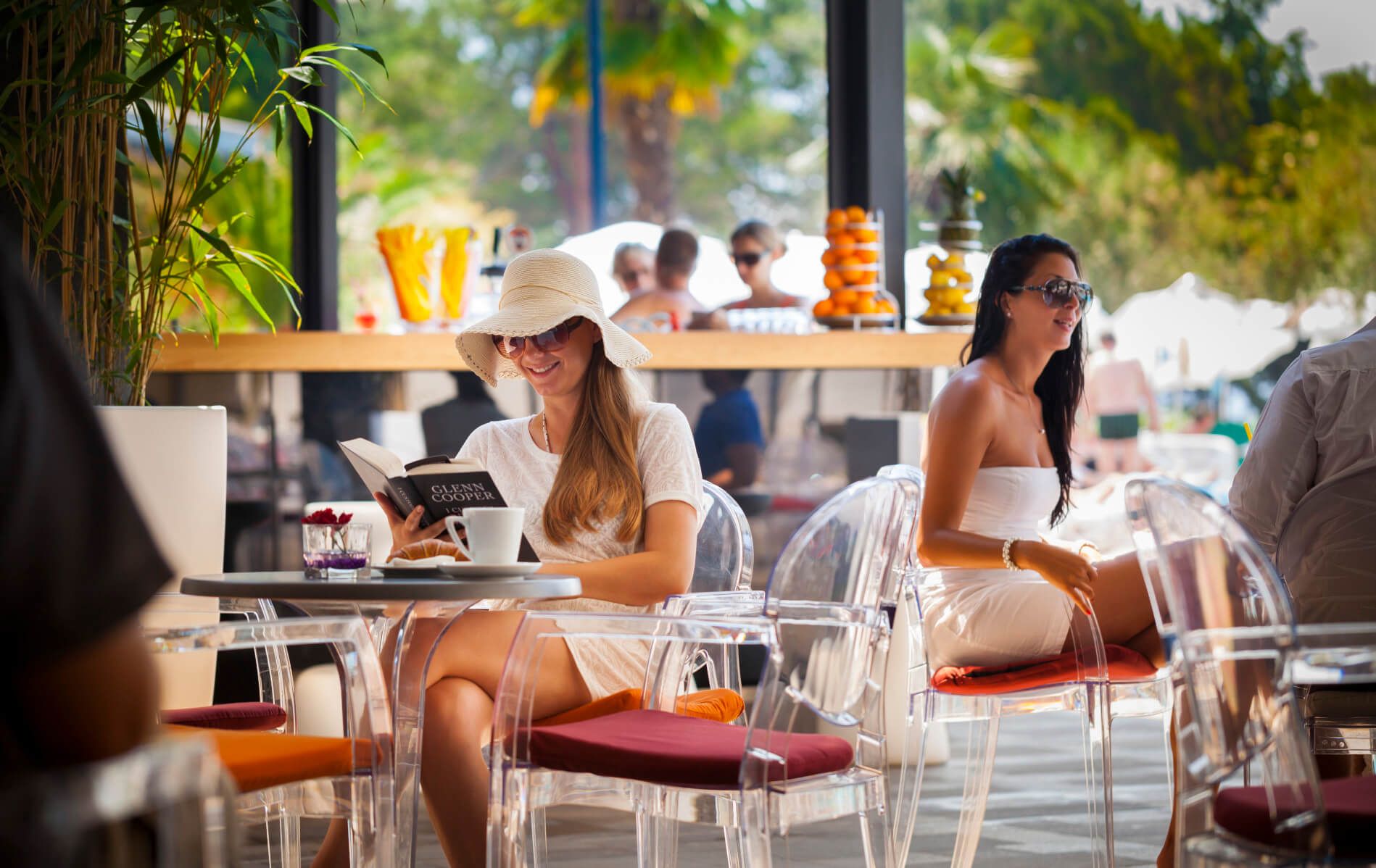 Eine Frau mit Hut liest in einem Café, im Hintergrund sind sonnige Gäste und Obstständer zu sehen.