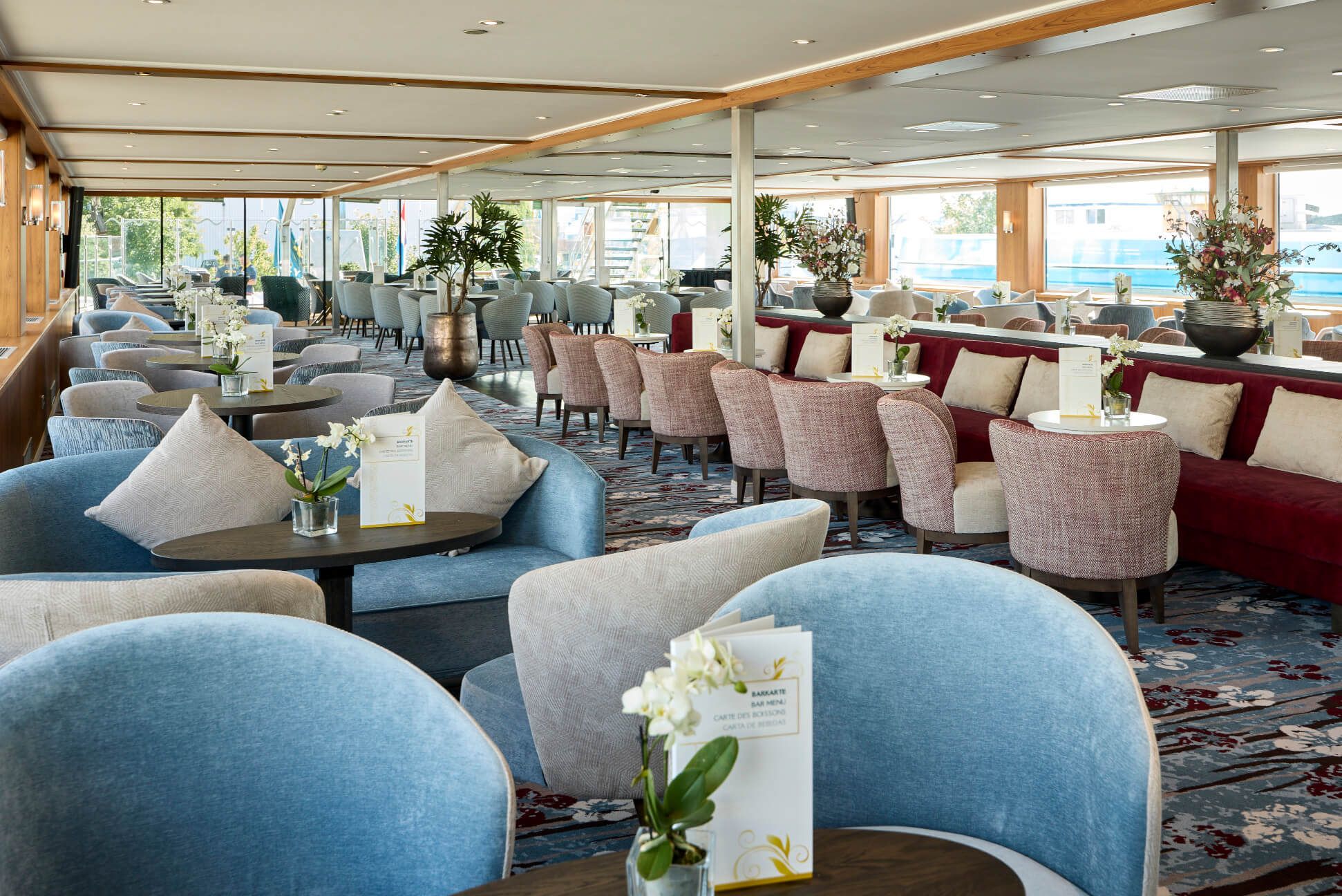 Eleganter Lounge mit bequemen Sesseln, Tischen und Pflanzen, viel Licht durch große Fenster. Entspannte Atmosphäre.