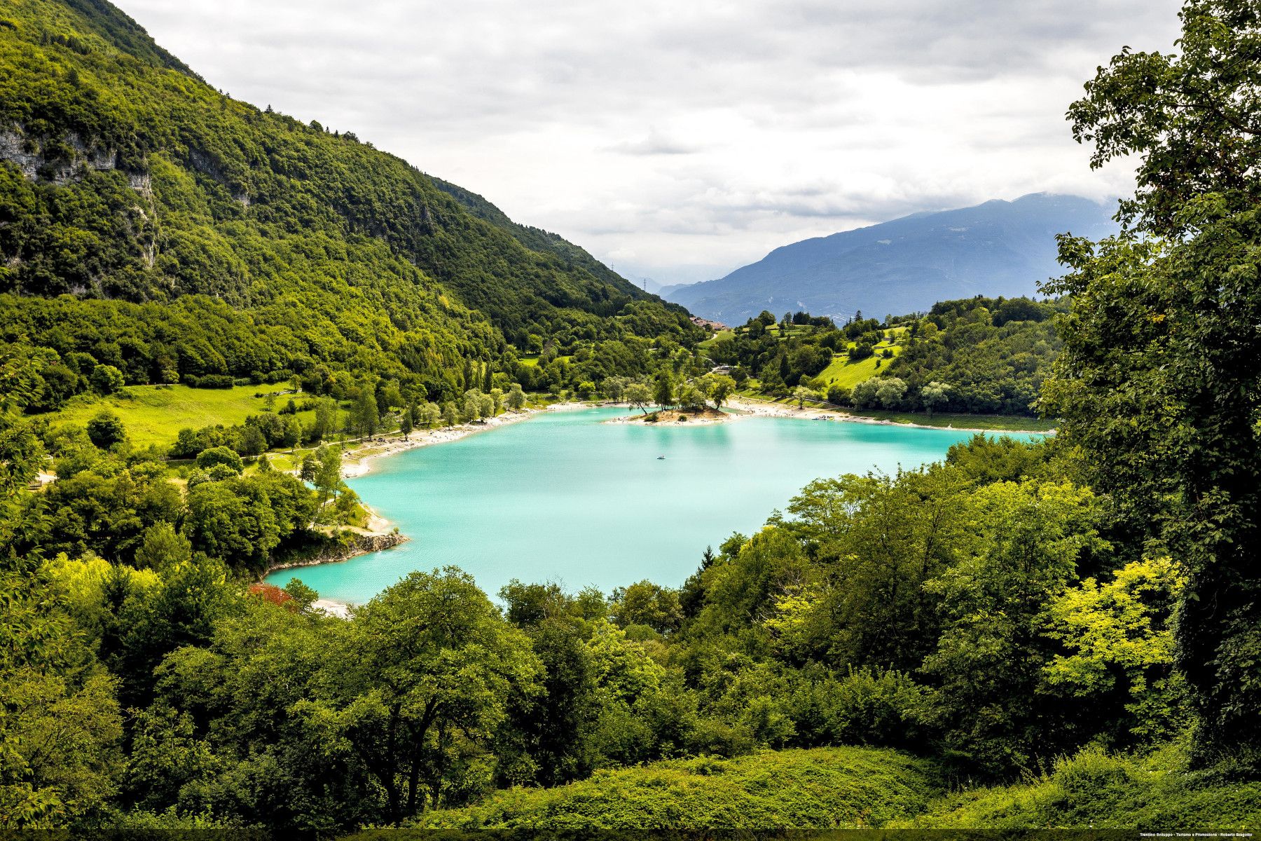 Eine malerische Landschaft mit einem blauen See, umgeben von grünen Bergen und Wäldern unter einem bewölkten Himmel.