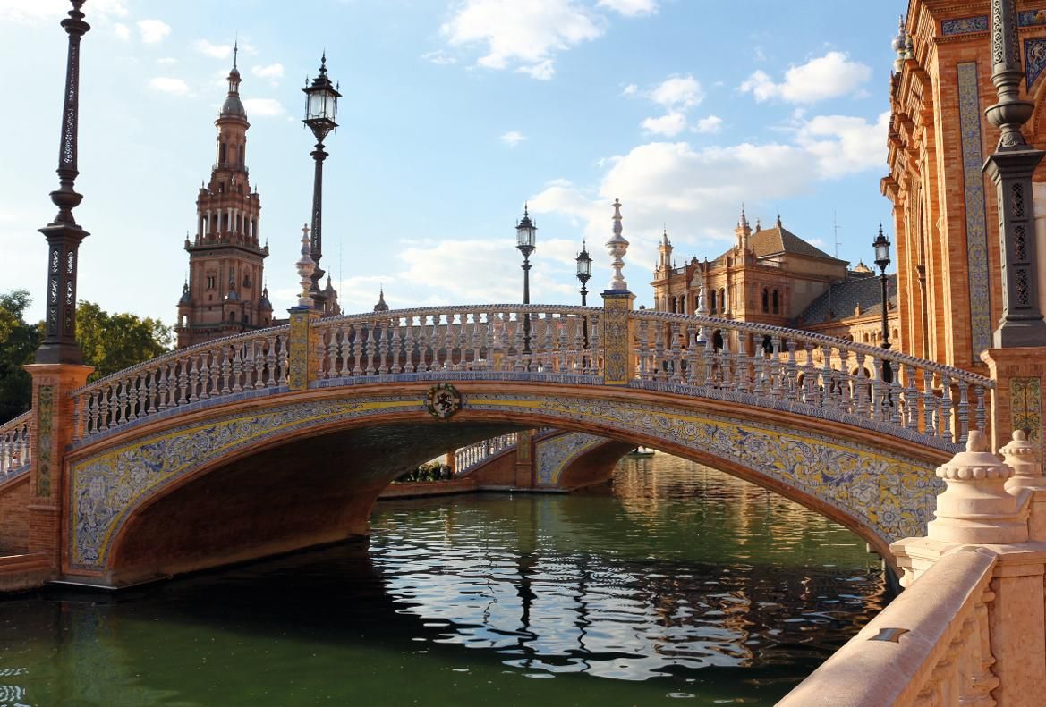 Eine kunstvolle Brücke über ruhiges Wasser, umgeben von beeindruckender Architektur und blauem Himmel.