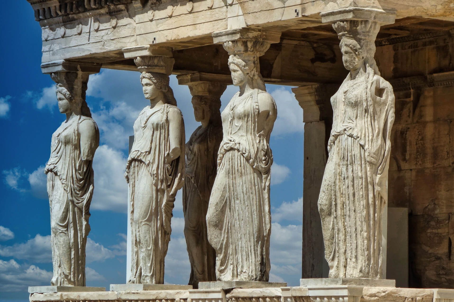 Die Abbildung zeigt die Karyatiden des Erechtheions, antike Statuen in Athen, vor einem blauen Himmel.