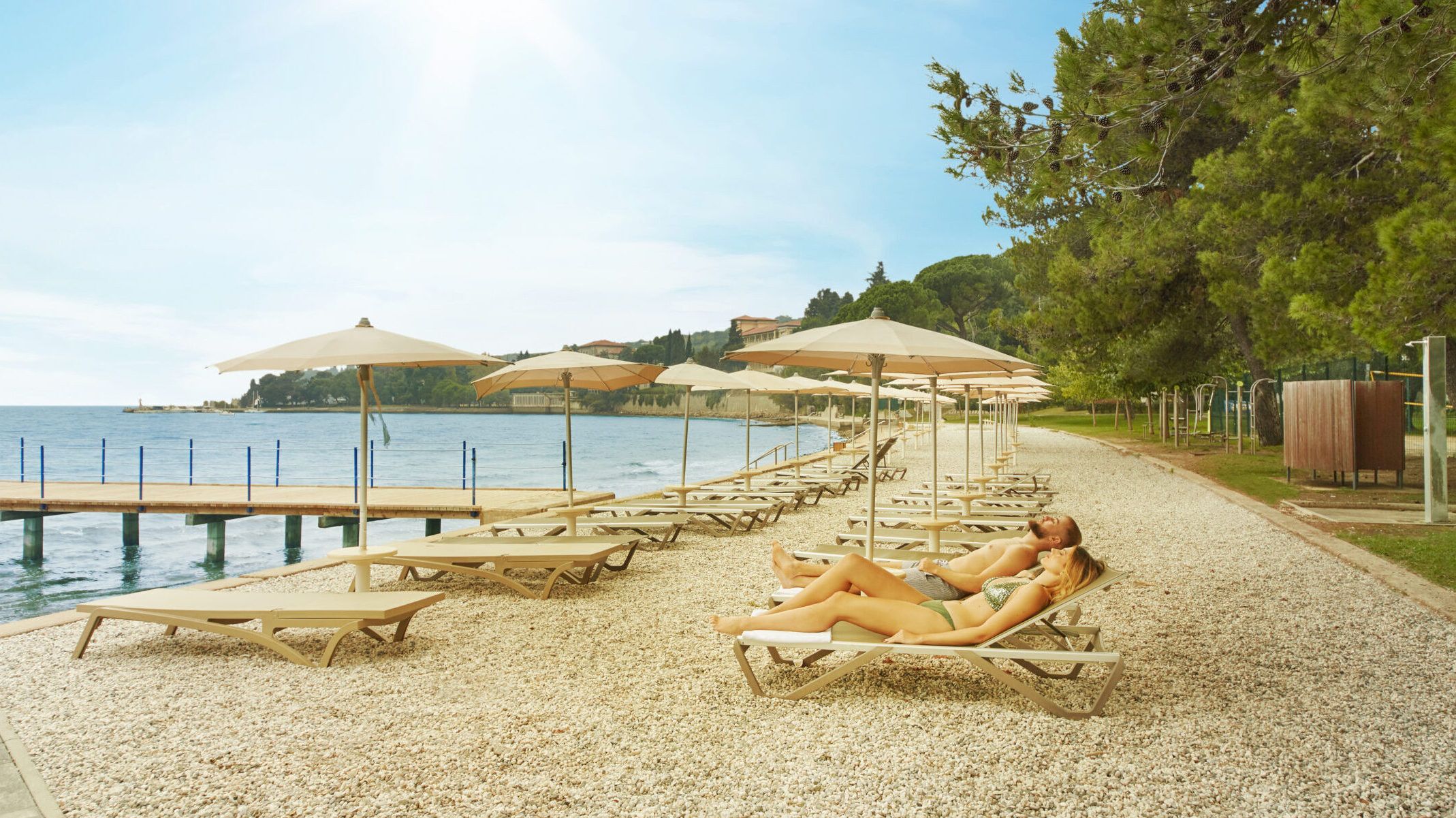 Ein sonniger Strand mit Liegen, Sonnenschirmen und zwei Badegästen, die sich am Wasser entspannen.