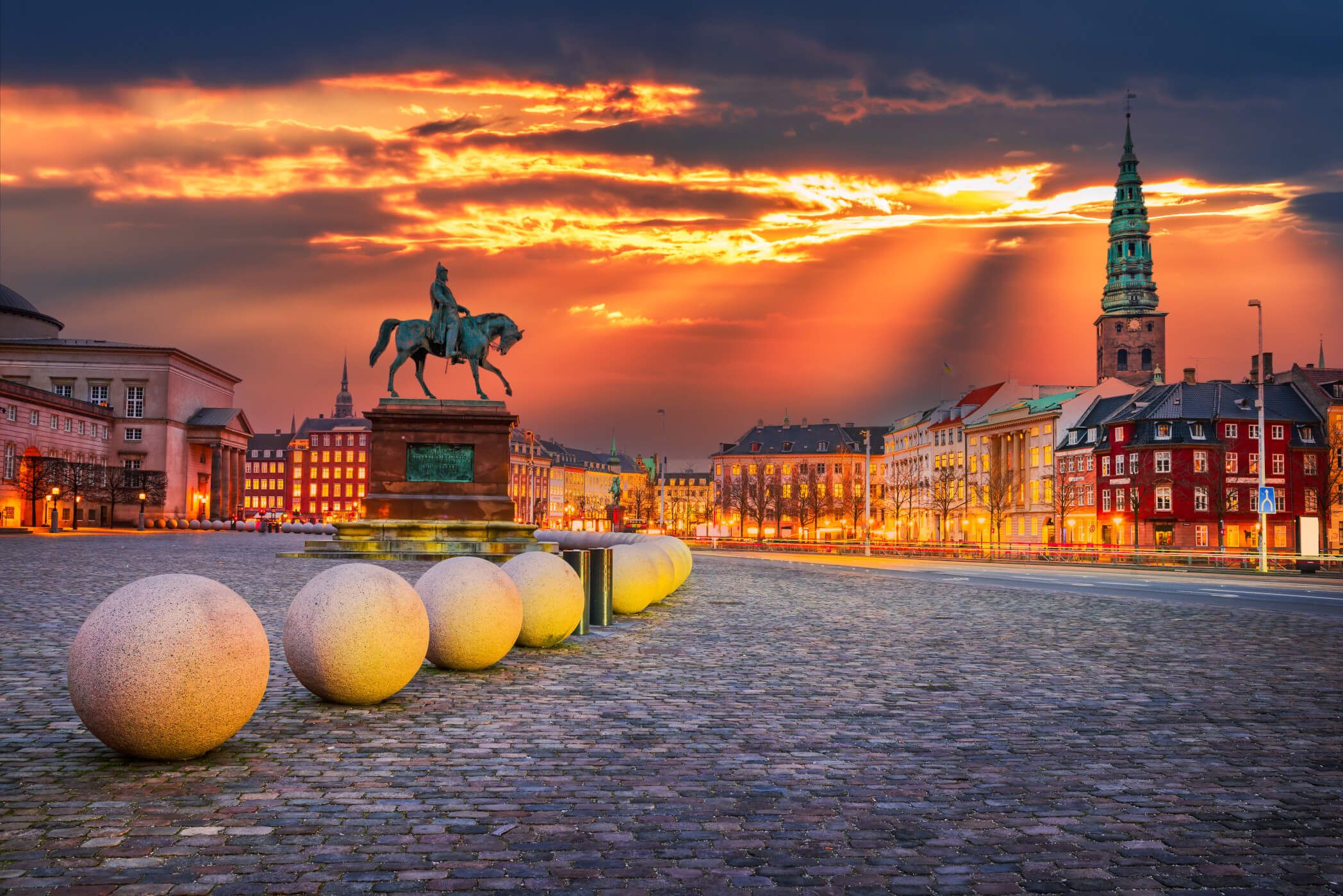 Ein Platz mit einem Reiterdenkmal, runden Steinen und bunten Gebäuden unter einem dramatischen Sonnenuntergang.