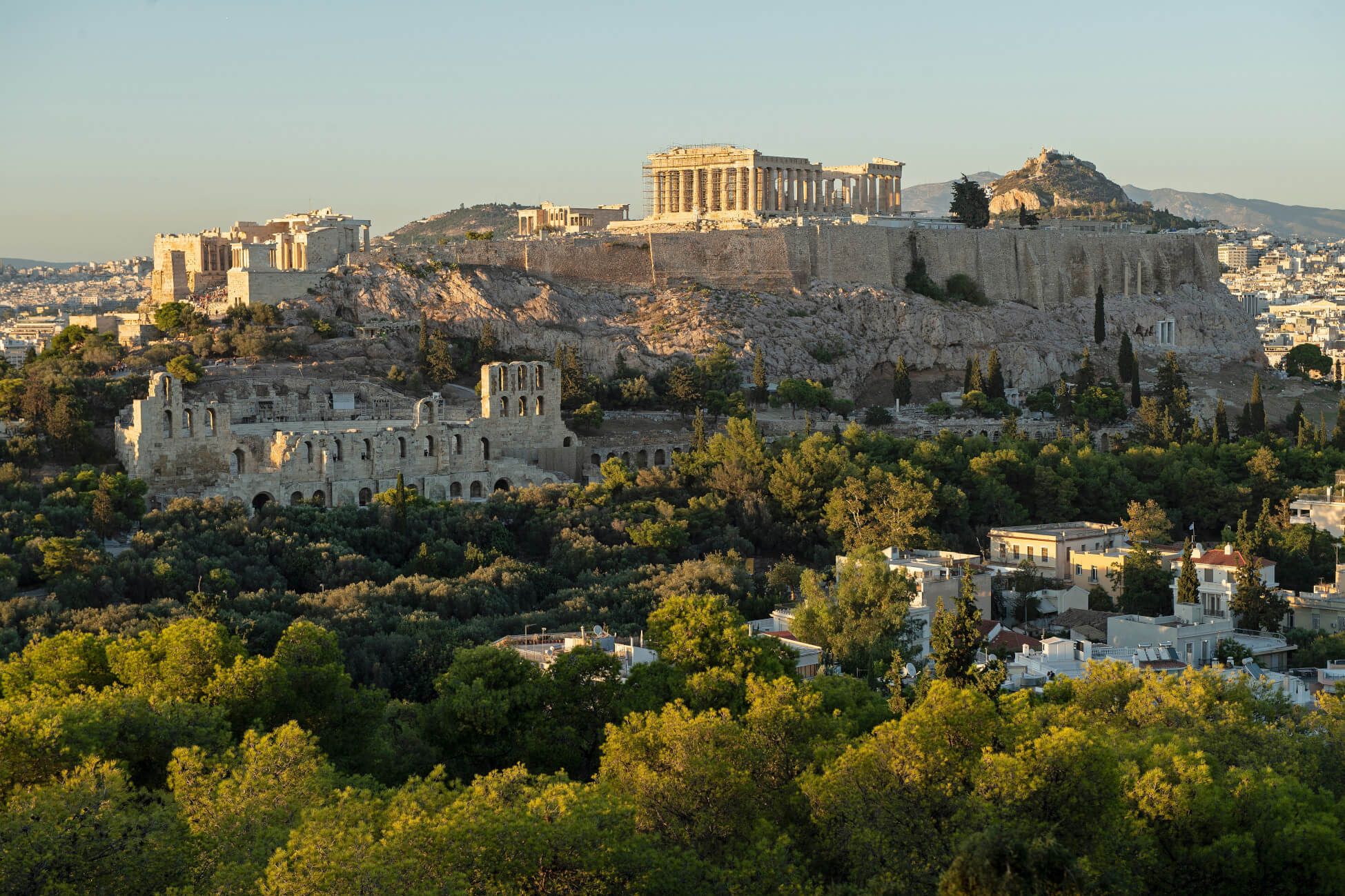 Athen mit der Akropolis und antiken Ruinen, umgeben von üppigem Grün und moderner Architektur im Vordergrund.