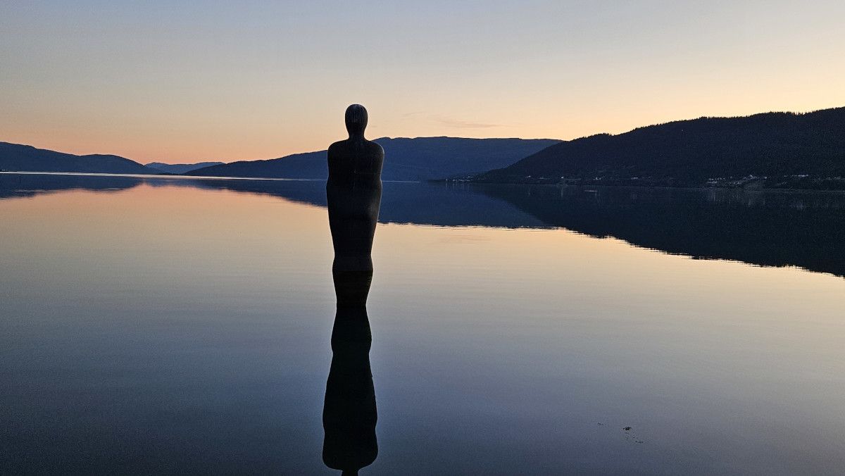 Eine Statue steht im Wasser, umgeben von ruhigen Bergen und einem sanften, farbenfrohen Sonnenuntergang.
