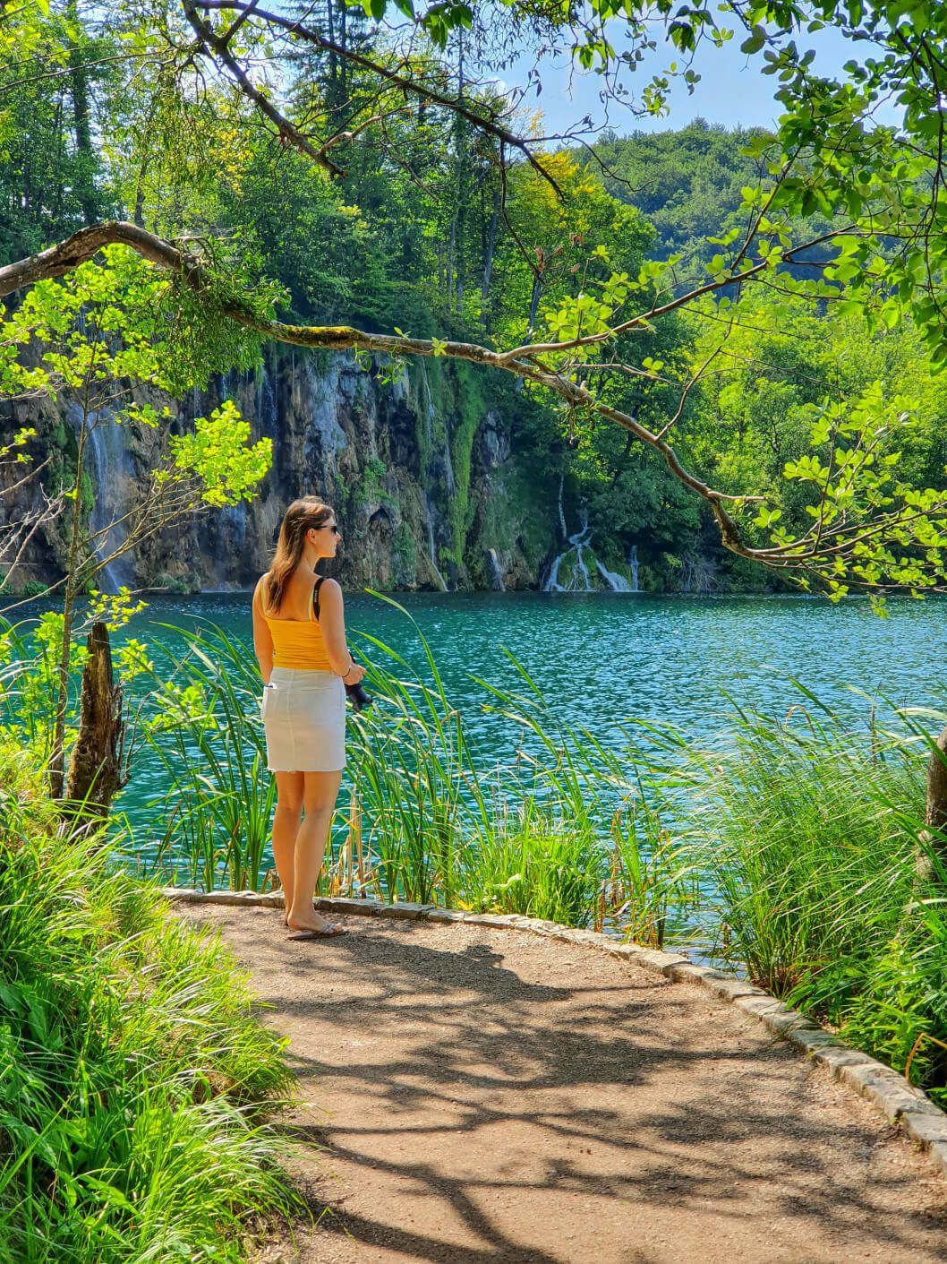 Eine Frau mit braunen Haaren steht am Wasser, umgeben von Bäumen und grünem Ufer, an einem sonnigen Tag.