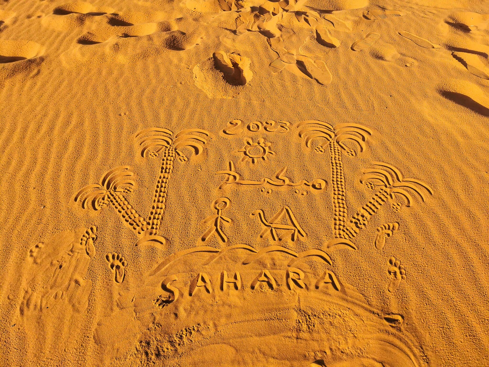 Ein Sandbild im Sahara mit Palmen, Sonne und den Jahren 2023. Fußabdrücke sind ebenfalls sichtbar.