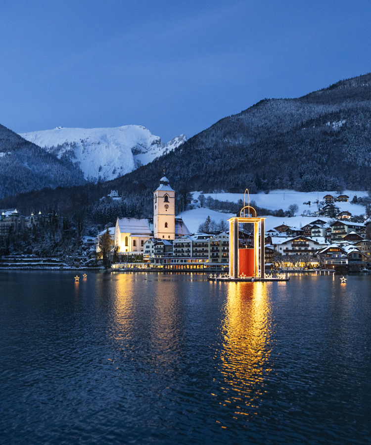 Ein winterliches Dorf am See bei Dämmerung, mit schneebedeckten Bergen und leuchtenden Elementen im Wasser.