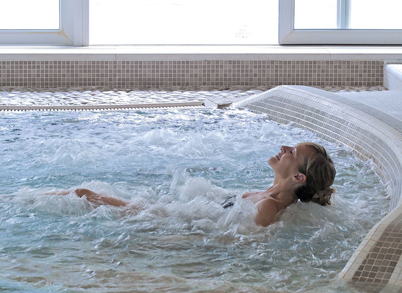 Eine Frau entspannt im Sprudelbad und genießt das Wasser, umgeben von Licht und einer ruhigen Atmosphäre.
