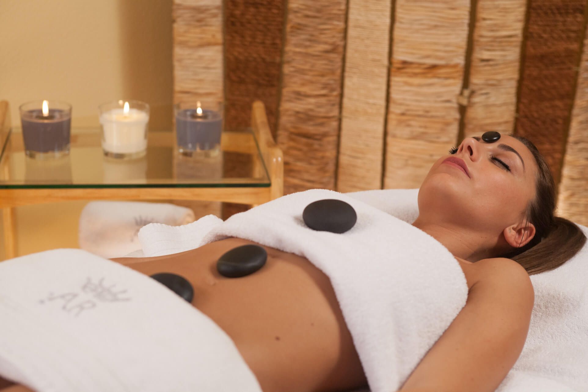 Eine Frau liegt entspannt auf einer Liege, mit heißen Steinen und Kerzen in einem Wellnessbereich.