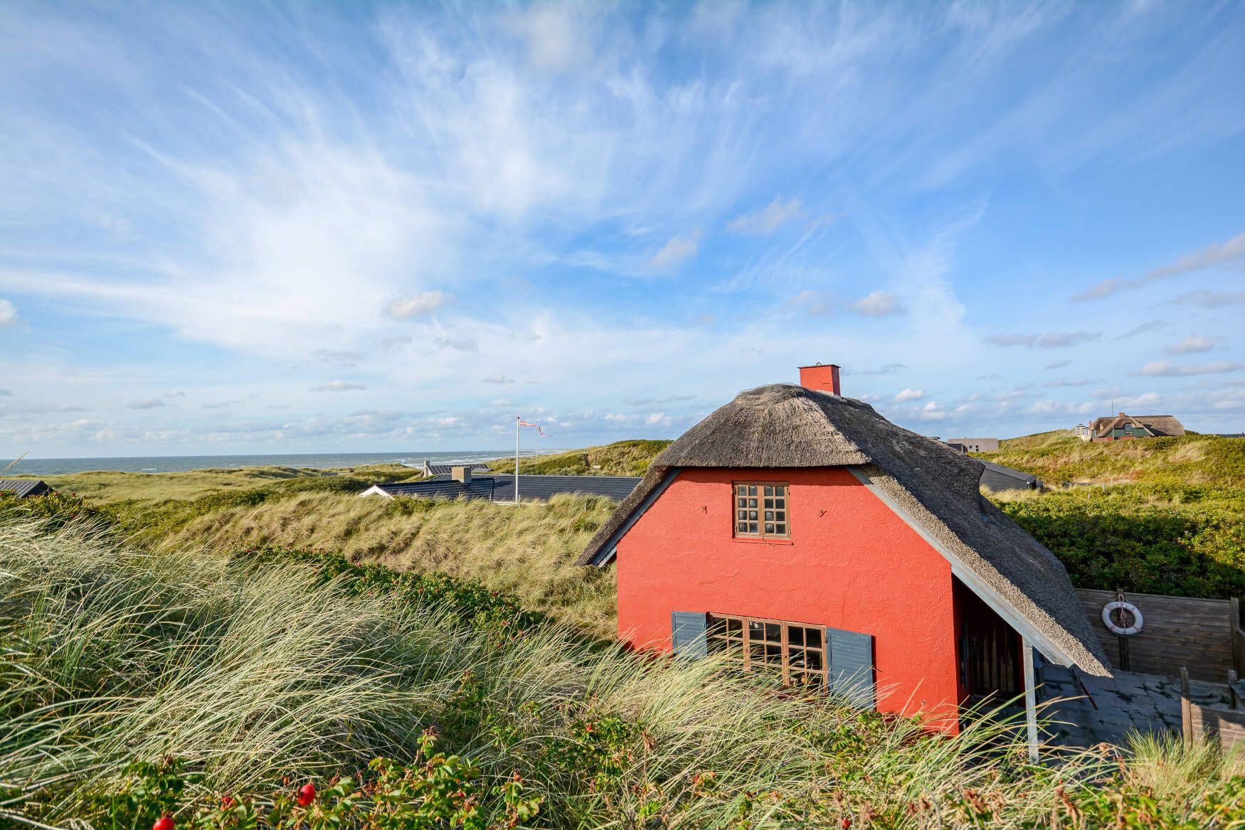 Ein rotes Haus mit Reetdach in einer Dünenlandschaft, umgeben von Gras und blühenden Pflanzen, unter blauem Himmel.
