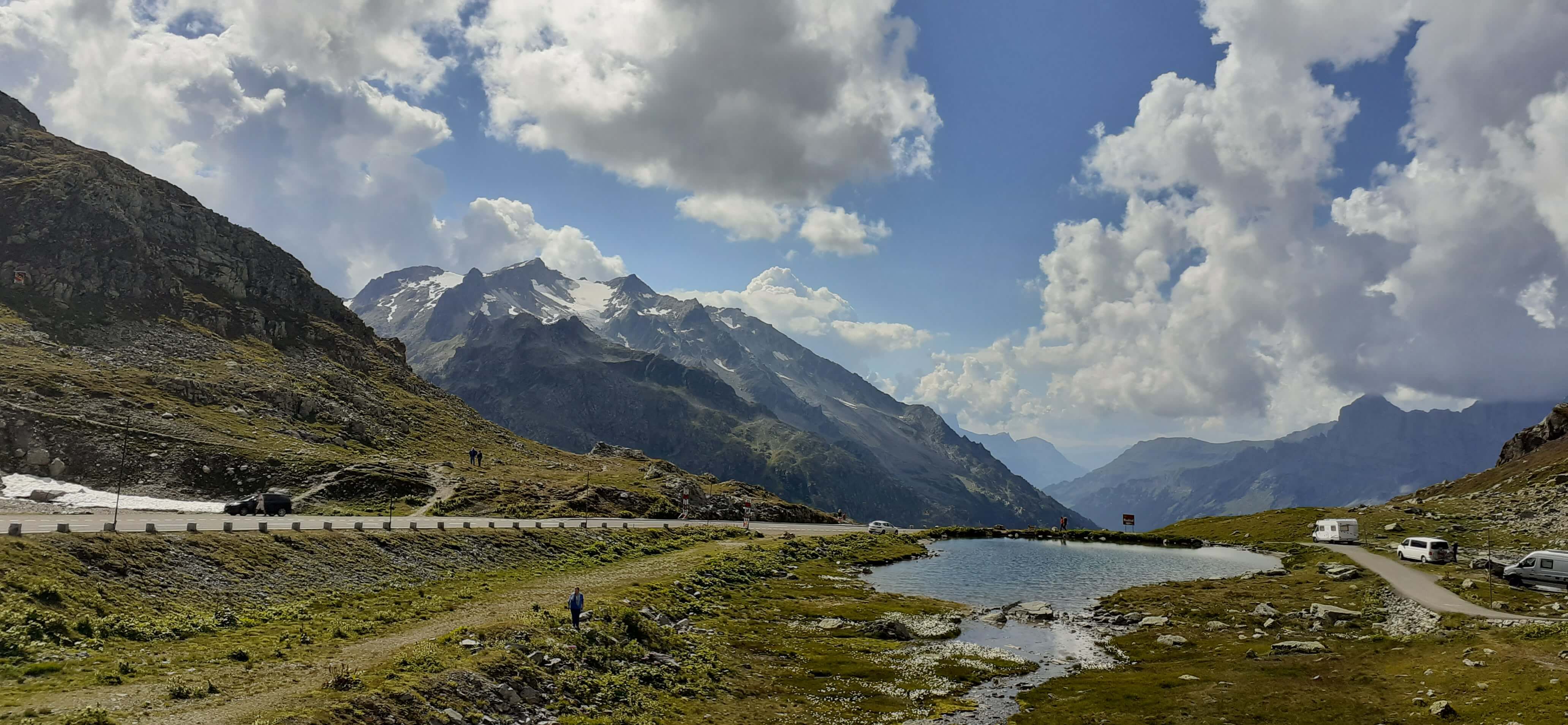 Eine malerische Berglandschaft mit Schneegipfeln, Wolken und einem klaren See, umgeben von grünen Wiesen.