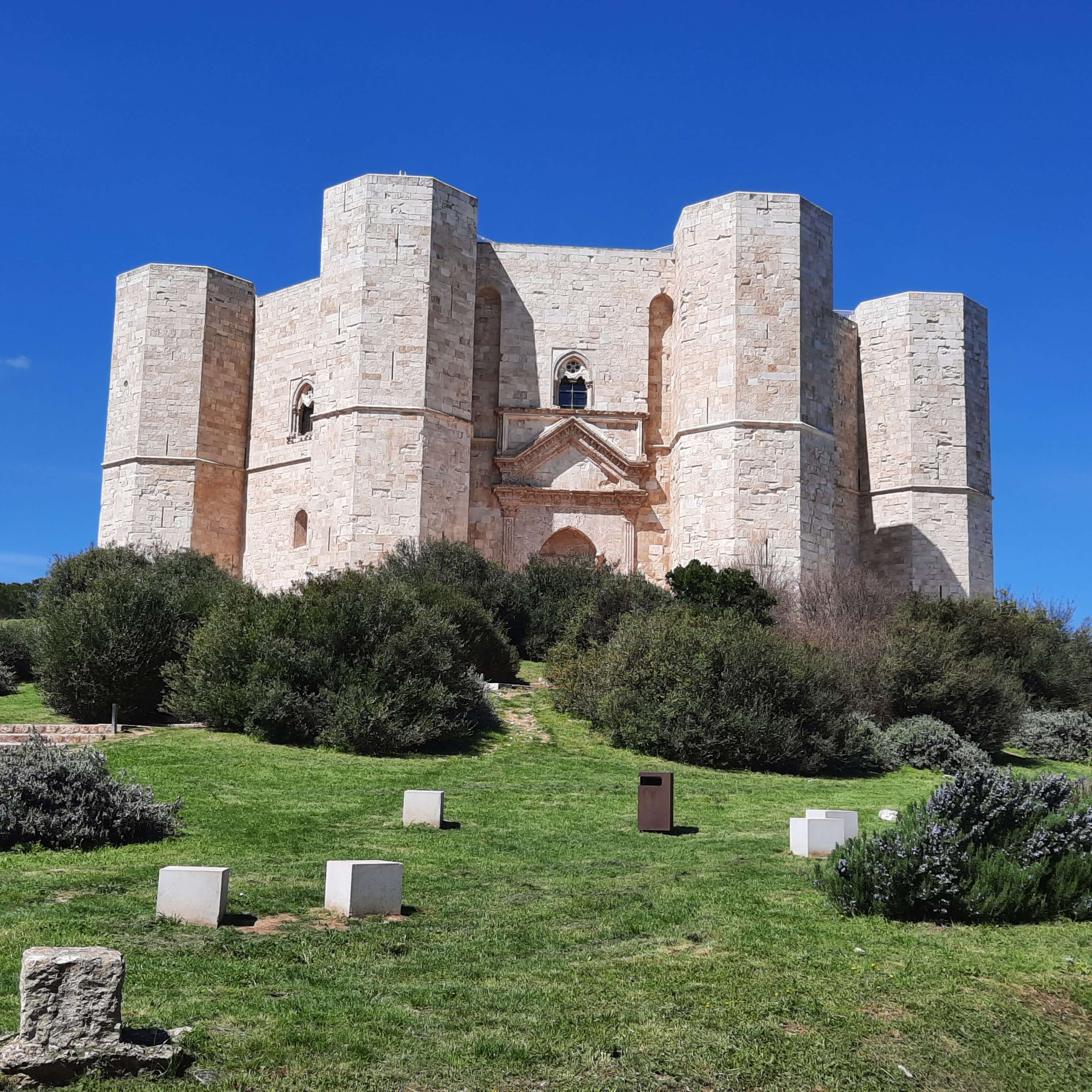 Eine beeindruckende Burg mit imposanten Türmen, umgeben von grünen Sträuchern und blauem Himmel.