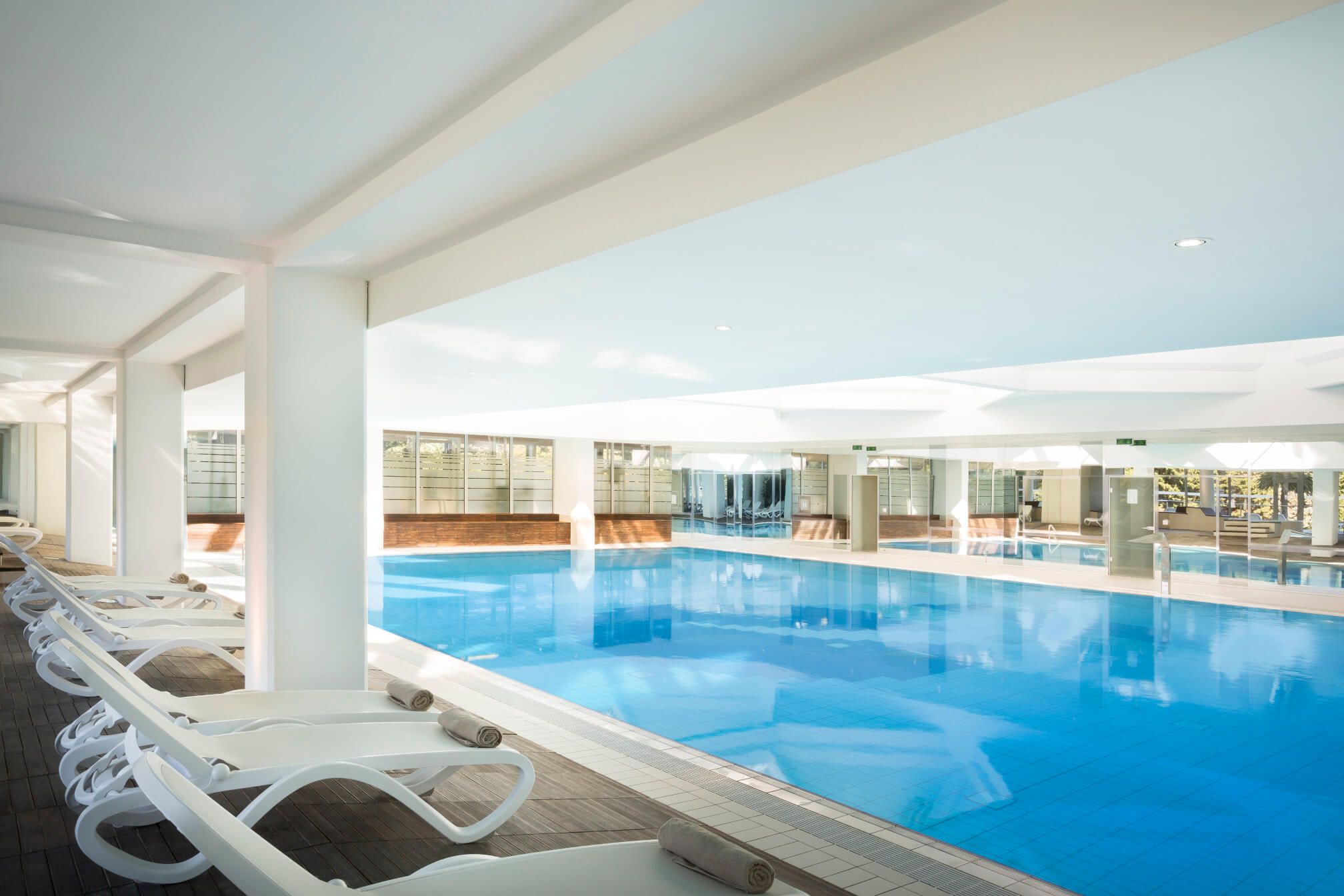Ein helles Hallenbad mit modernem Design und Liegen zum Entspannen am Rand. Klarer blauer Wasser reflektiert Licht.