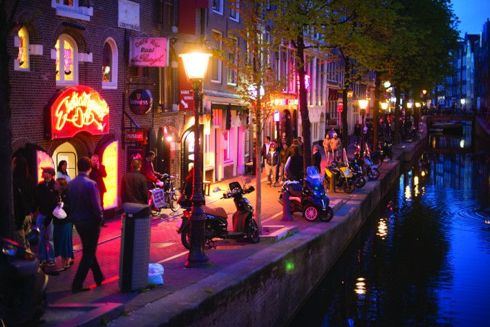 Bunte Lichter erhellen die Amsterdamer Straße, Menschen und Motorräder am Kanal, abendliche Atmosphäre.