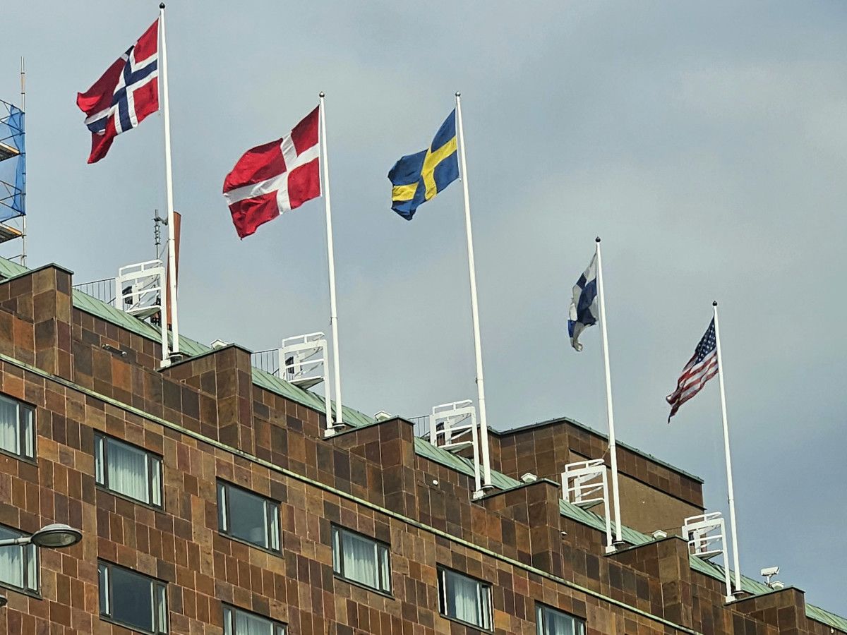 Fünf Flaggen wehen über einem Gebäude: Norwegen, Dänemark, Schweden, Finnland und die USA. Der Himmel ist bewölkt.
