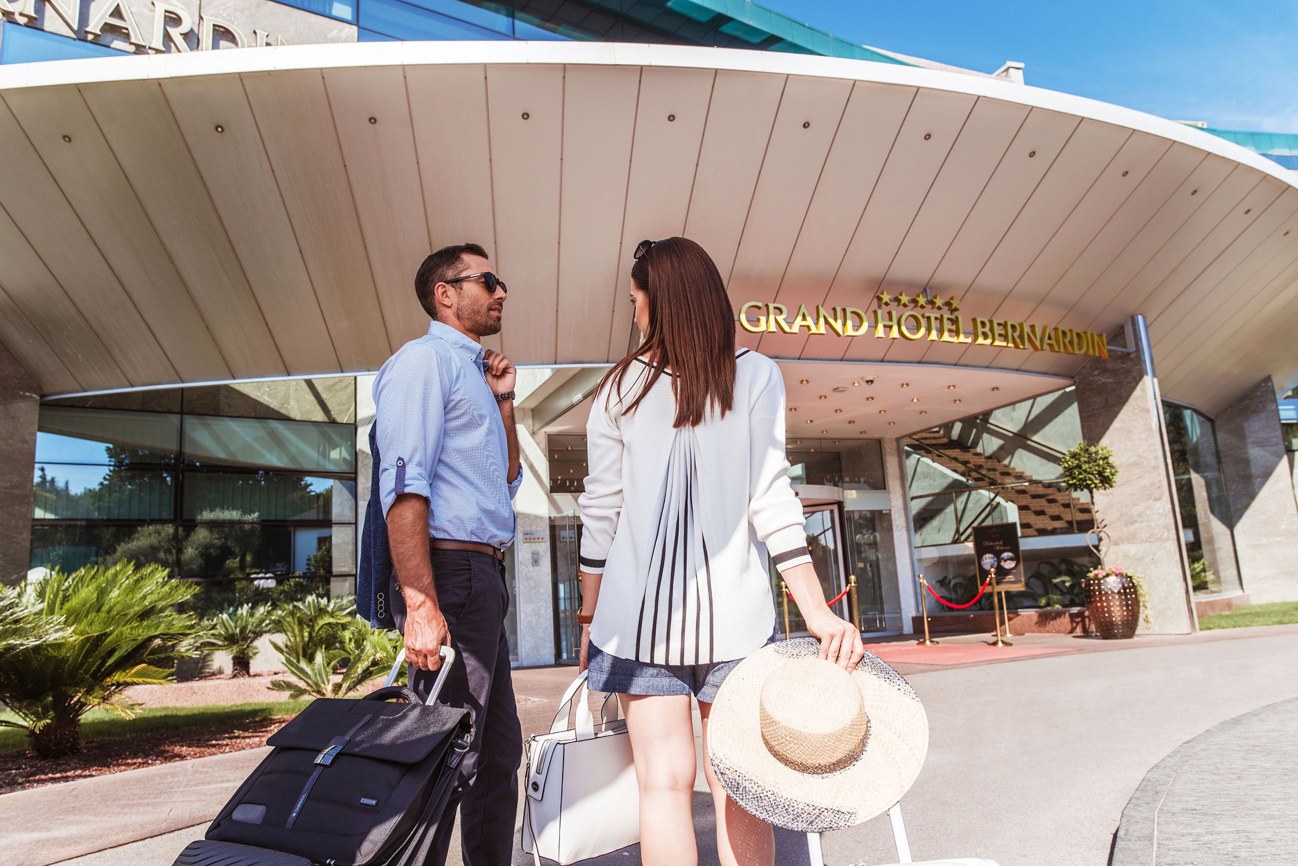 Paar vor dem Grand Hotel Bernardin, bereit für den Check-in mit Gepäck und Sonnenhut. Sonniger Tag, entspannte Stimmung.